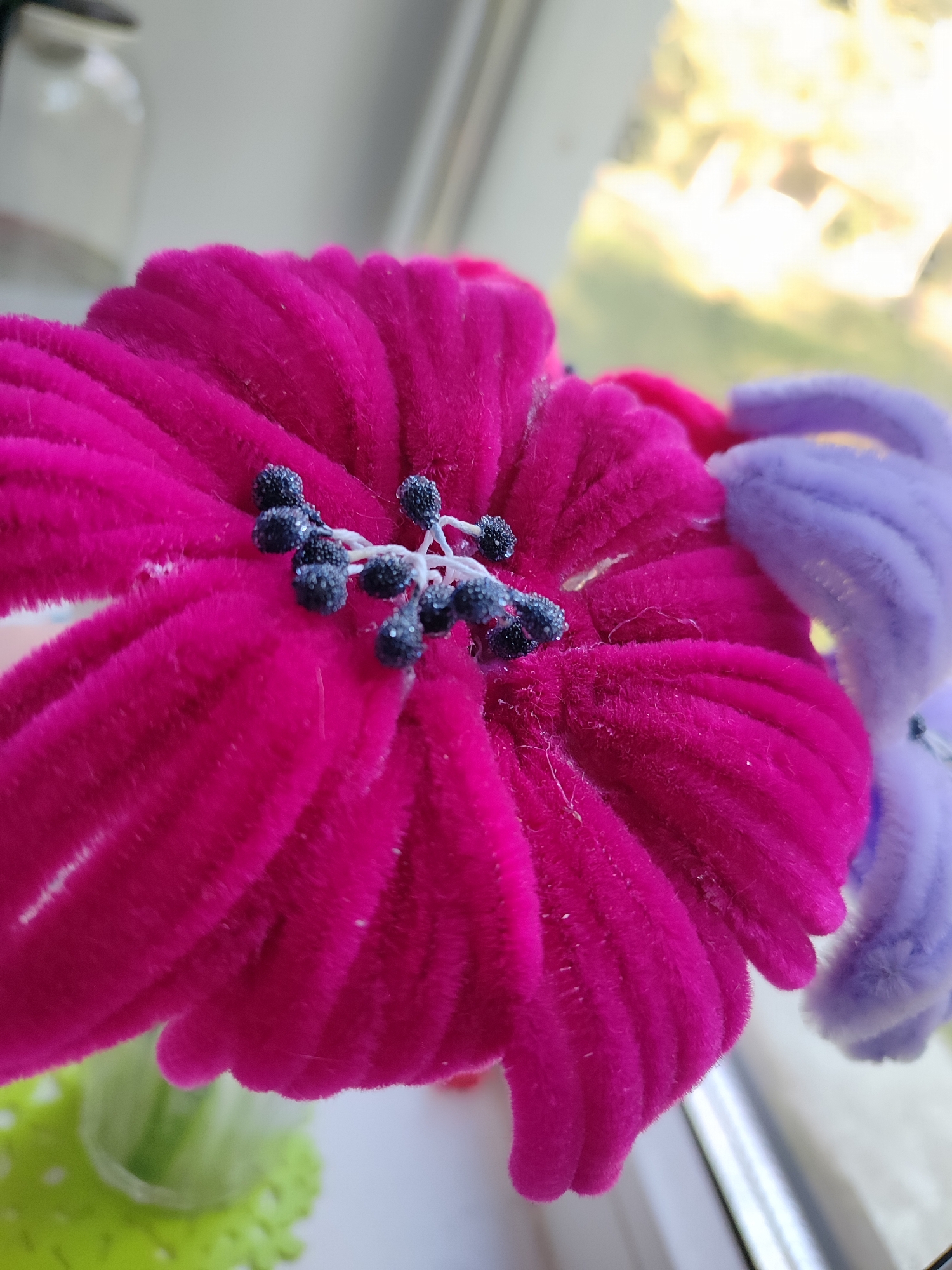 A close-up, eye-level, outdoor shot of two artificial flowers made of pipe cleaners is shown. The flower in the foreground is bright pink, and the flower in the background is light purple. The pink flower has a center of dark beads on a white filament. The background is blurred and shows trees and foliage through a window.