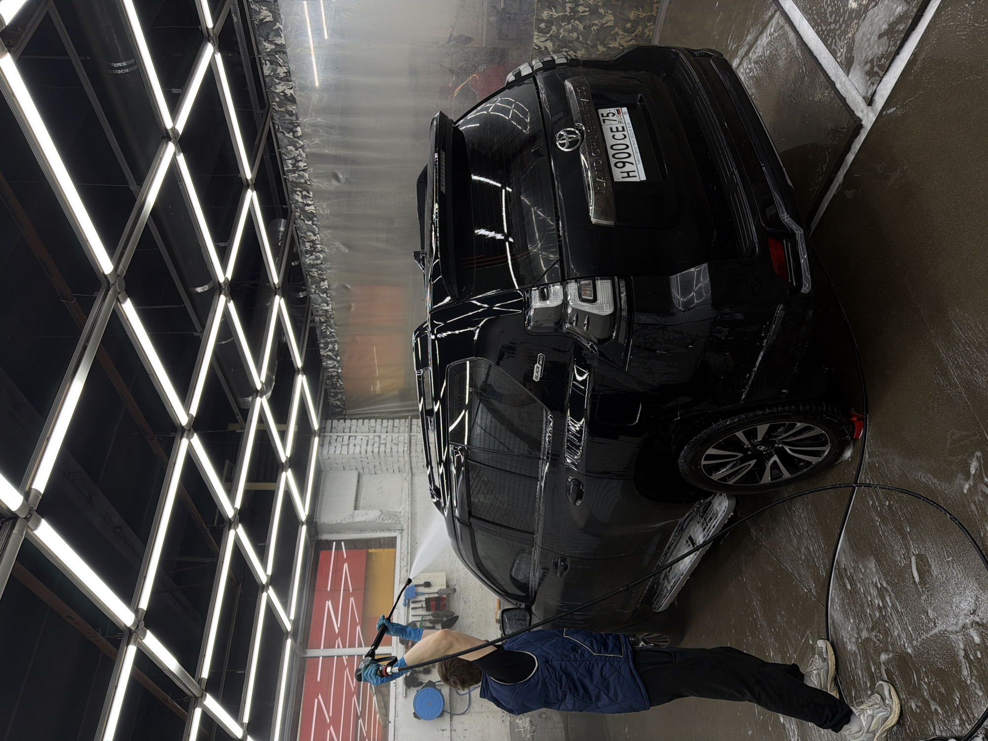 A black Toyota Land Cruiser Prado SUV with license plate "H 900 CE 75 RUS" is undergoing a car wash at an indoor location. A person in blue gloves, a blue vest, black pants, and gray sneakers is seen washing the car with a high-pressure water hose. The location appears to be in Krasnoyarsk, Russia, based on the license plate region code. The background includes a wall of fluorescent lights arranged in a crisscross pattern and a wall with a red and yellow geometric design. The floor is wet and covered in soapy water.