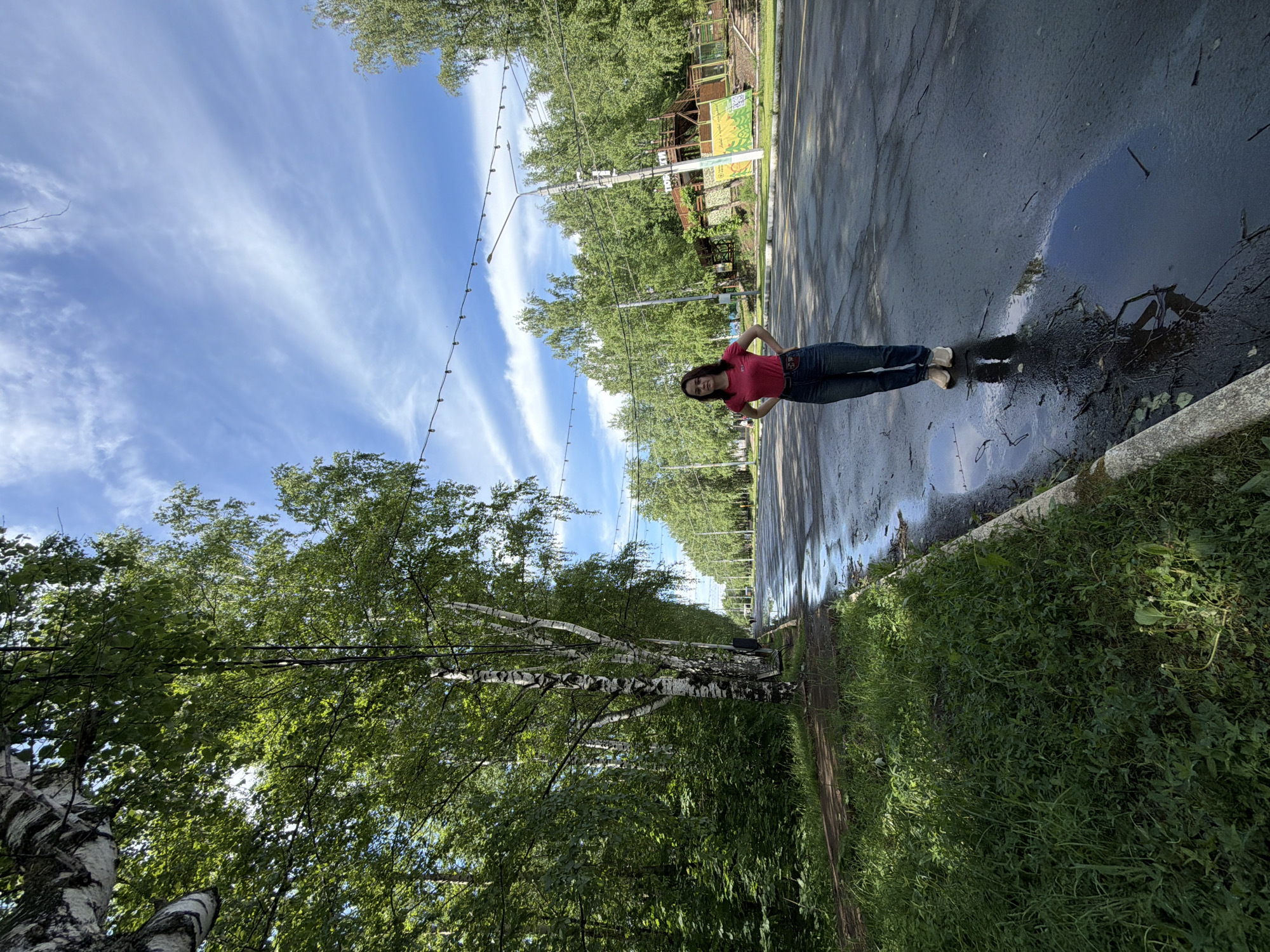 A woman stands on a wet road in Izhevsk, Russia. She wears a red shirt and blue jeans. Her hands are on her hips. A puddle near the curb reflects the sky. Trees line the road, and overhead string lights are visible. A wooden structure stands on the opposite side of the road. The day appears sunny with a partly cloudy sky. A grassy area is beside the curb. Birch trees are visible. The road is wet.
