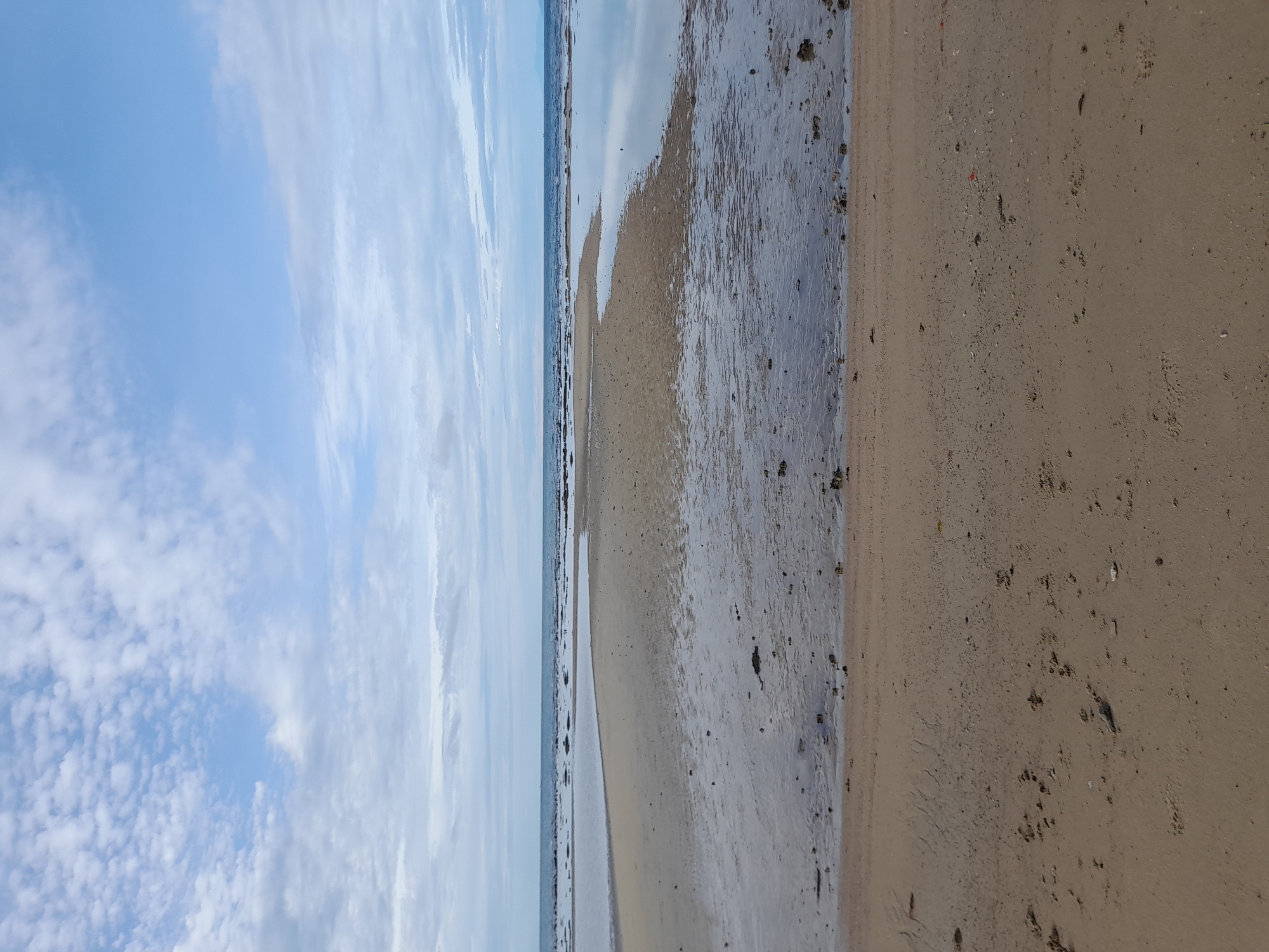 The image shows a beach scene with sand in the foreground and shallow water with exposed sandbanks in the mid-ground. The sky is partly cloudy with a mix of white and gray clouds against a pale blue sky. The horizon is visible where the sea meets the sky, with a darker strip of land or islands in the far distance. The overall lighting suggests it is daytime, likely overcast or with the sun not directly overhead. The scene is devoid of any people or structures.