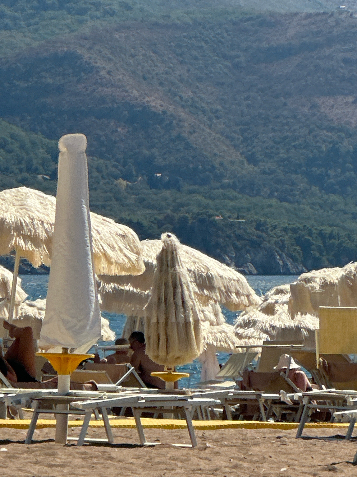 The image depicts a beach scene in Boreti, Montenegro, on a sunny day. In the foreground, several lounge chairs are set up on the sand, some occupied by people sunbathing. The chairs are mostly white or dark-colored, with some featuring yellow accents. Beach umbrellas, some with fringed canopies and others with plain fabric, provide shade. The umbrellas are a mix of cream, beige, and white colors.
In the middle ground, the turquoise water of the sea is visible, with gentle waves lapping at the shore. The background is dominated by a steep, green mountainside covered in dense vegetation, with rocky outcrops visible in places. The sky is clear and blue.
One person is clearly visible in a lounge chair, wearing a light-colored hat and swimwear, facing away from the camera. Another person's legs are visible, lying on a lounge chair to the left. There are glimpses of other people under umbrellas and on chairs, but they are not clearly distinguishable. The overall impression is one of a relaxed summer day at the beach.