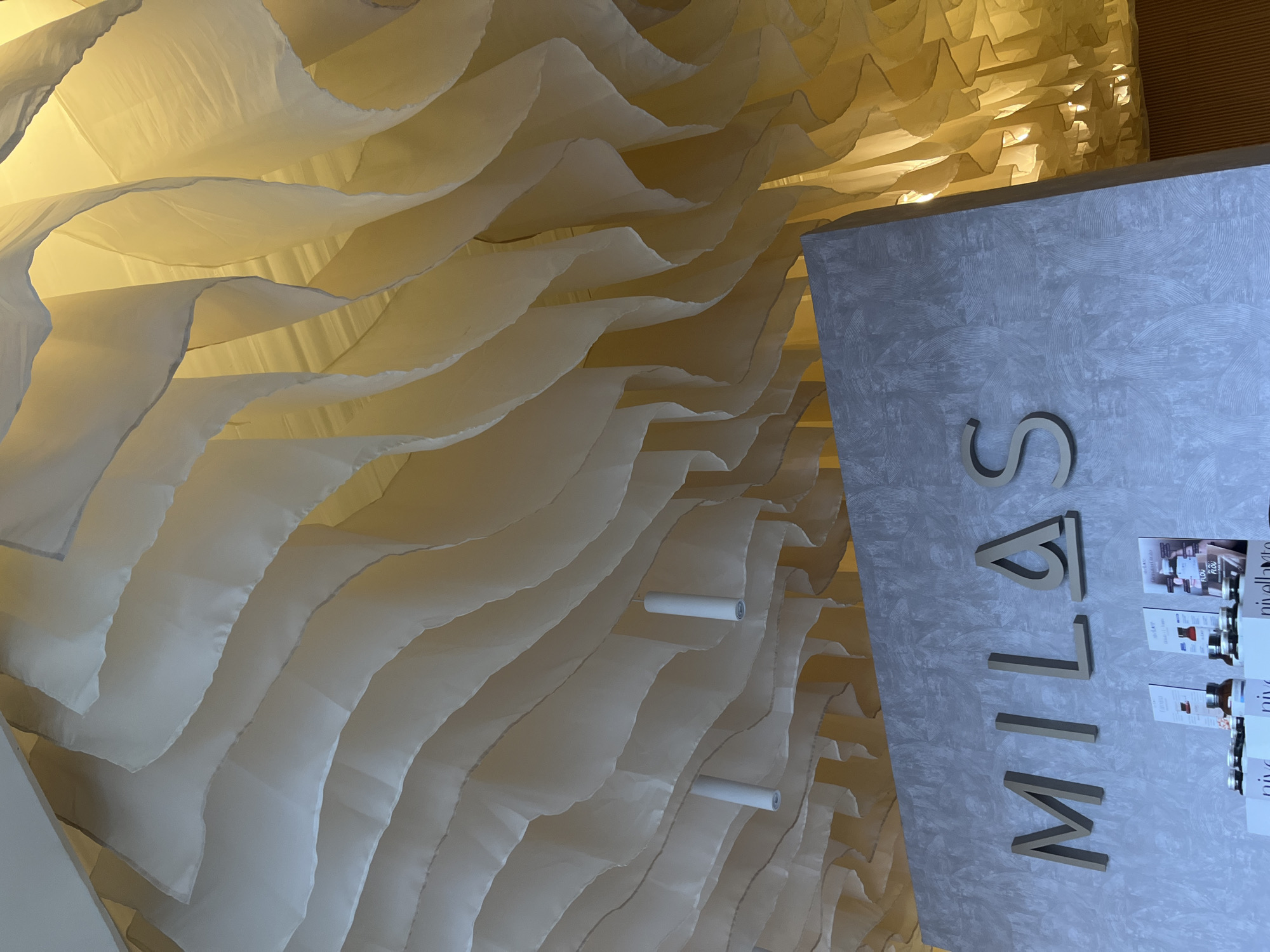 The image displays an interior view of a sophisticated commercial space, likely a store or salon, located in San Bernardino Tlaxcalancingo, Mexico. The dominant feature is an elaborate ceiling adorned with numerous layers of light-colored, semi-transparent fabric. These fabrics are artfully draped to create a soft, undulating, and wavy pattern, giving the impression of flowing textiles. Warm, golden-yellow light illuminates the fabric from behind or above, creating a glowing effect and highlighting the textures and contours of the drapes. Several white cylindrical objects, possibly lights or structural elements, are visible among the fabric layers.
On the right side of the frame, a grey wall with a subtle textured pattern of concentric circles or waves serves as a display area. Large, three-dimensional letters, in a golden or brass finish, are mounted on this wall, spelling out "MILAS". Below the "MILAS" sign, there is a display of small bottles and brochures, likely showcasing cosmetic or beauty products. Visible text on these items includes "nive", "nivellanto", and "FLOU FLOU".
No people or specific activities are present, allowing the focus to remain on the modern and elegant interior design. The warm lighting and refined decor evoke a sense of luxury and comfort, suggesting an upscale establishment.