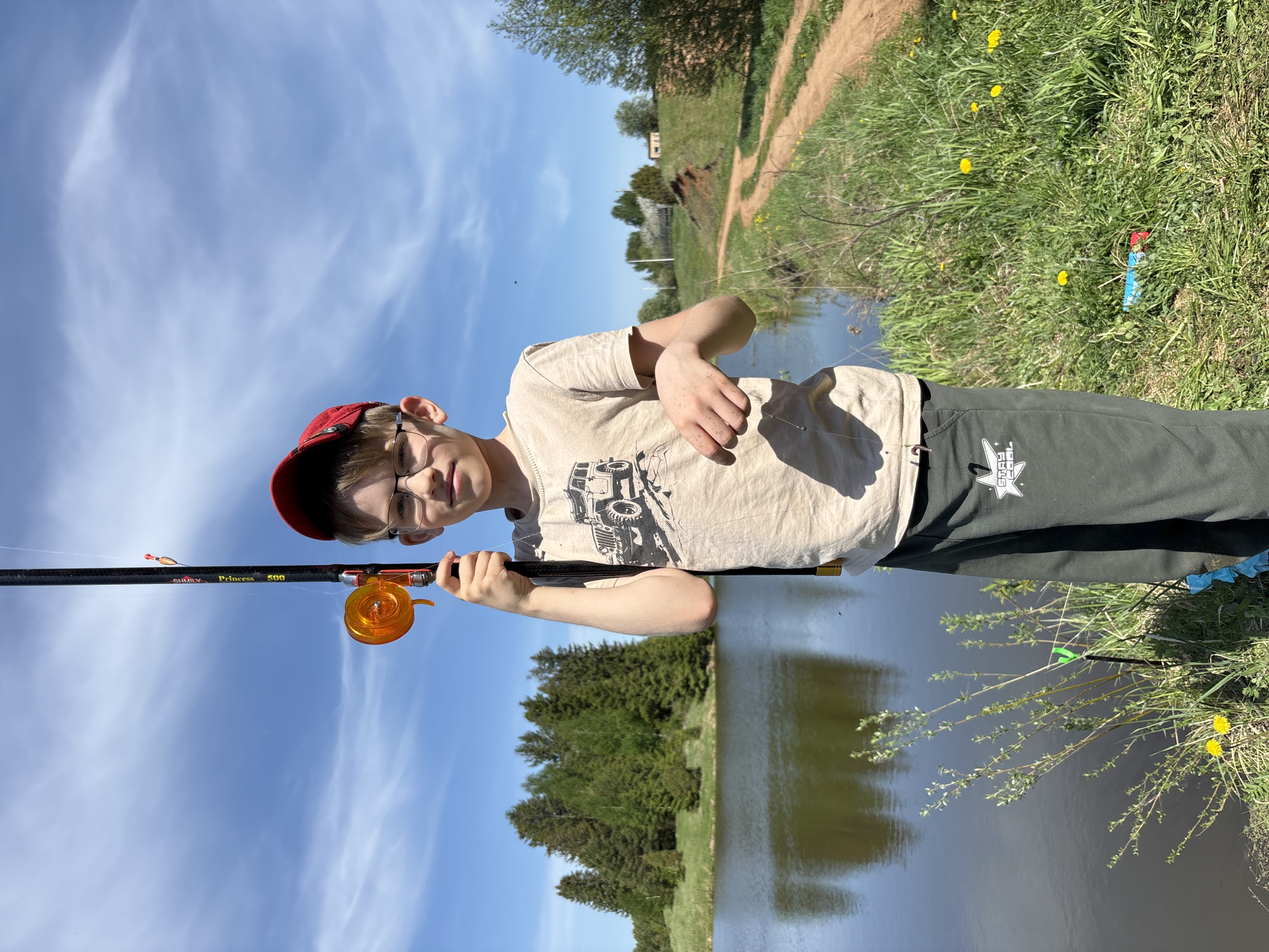 A young boy wearing a red cap and glasses stands at the edge of a lake. He holds a fishing rod with an orange reel. The rod has the name "Princess 500" written on it. The boy is wearing a light-colored t-shirt with a graphic on it and dark-colored pants with a white logo. The lake is surrounded by green grass and some yellow dandelions. A dirt path is visible in the background. The sky is blue with some clouds. The location is most likely Karashur, Russia.
