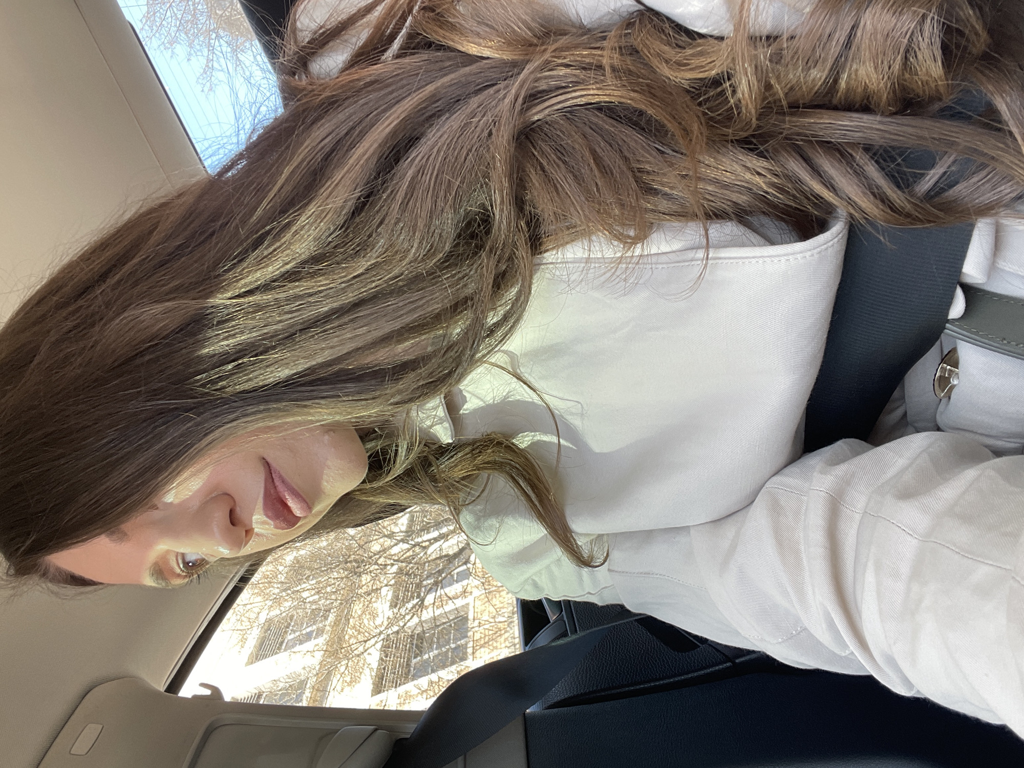 A woman with long brown hair is sitting in a car. She is wearing a light-colored hooded jacket and a seatbelt. Outside the car window, a building with many windows and bare trees is visible. The location is not discernible as Varna, Bulgaria.
