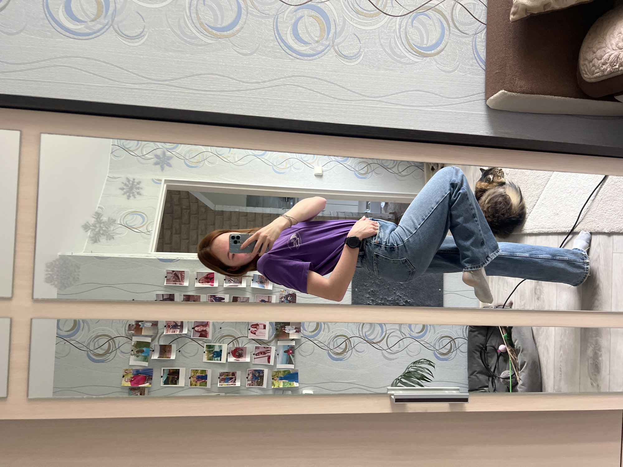 A young woman with reddish-brown hair is taking a selfie in a multi-paneled mirror. She is wearing a purple t-shirt and jeans. A cat is visible in the background on a rug. On the wall behind her reflection are several square photos arranged in a grid pattern. In the reflection, a door is partially visible in the background. There is a green plant to the right. The location is Chaikovsky, Russia.