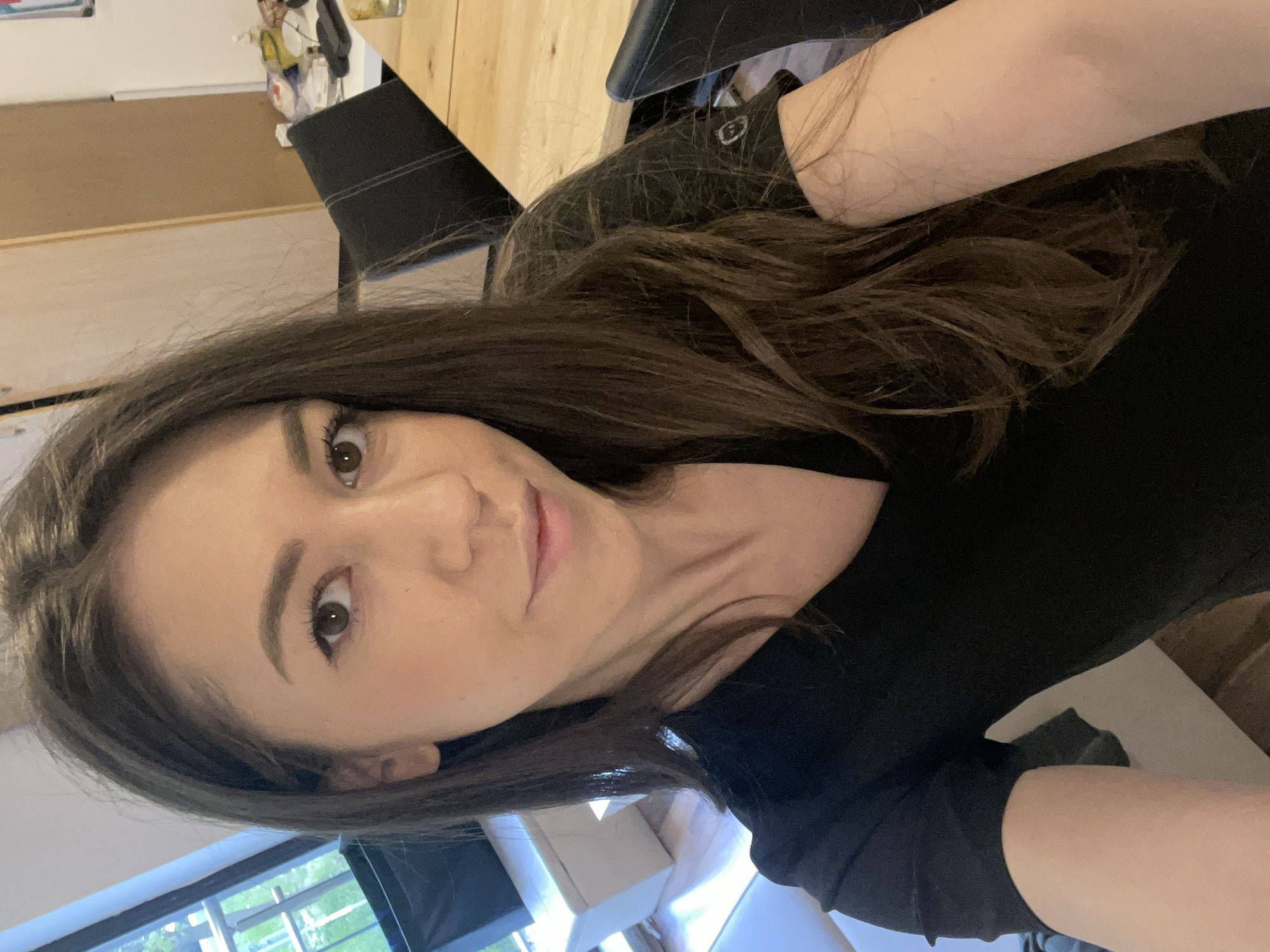 A woman with brown hair and eyes is centered in the frame, wearing a black shirt. Her head is tilted slightly, with her gaze directed towards the viewer. She appears to be taking a selfie. The background includes a window with a view of trees and grass, suggesting an outdoor setting. A light-colored sofa is visible behind her. To the left and behind her head there is a glimpse of a wooden table and chair. The location is likely indoors, possibly a home or apartment in Sofia, Bulgaria, based on the assumption this is her location.