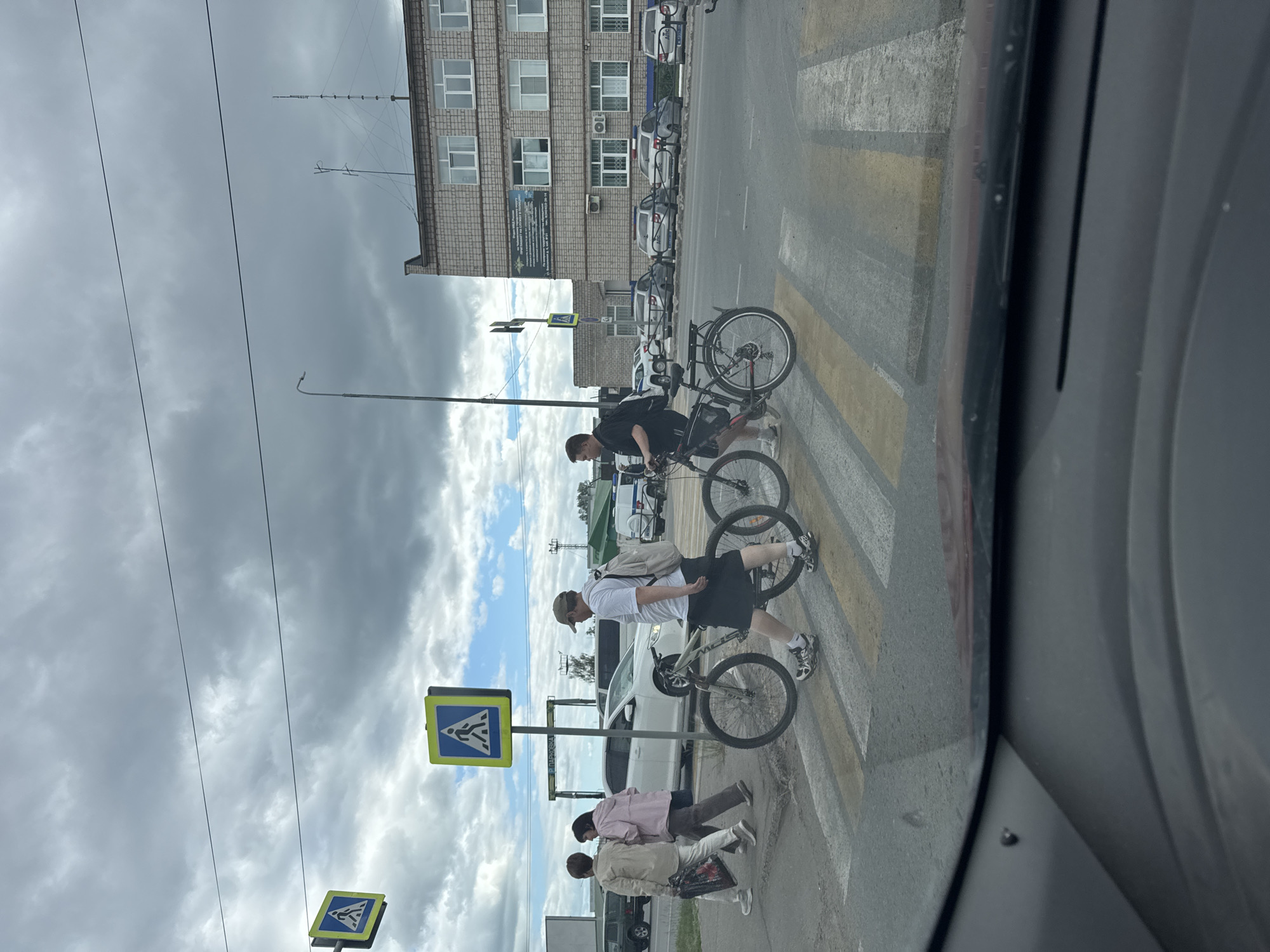In Chaikovsky, Russia, on a cloudy day, pedestrians and cyclists navigate a crosswalk in front of a brick building. Two individuals are pushing bicycles across the yellow and white striped crosswalk. To the left, two women walk across the crosswalk, one carrying a bag. Behind them, a white car waits at the intersection. A pedestrian crossing sign is visible to the left of the crosswalk. The brick building has multiple windows and signs, including what appears to be official signage. In the background, several cars, including some police vehicles, are parked along the street. The image is taken from inside a vehicle, as indicated by the car's dashboard in the foreground.