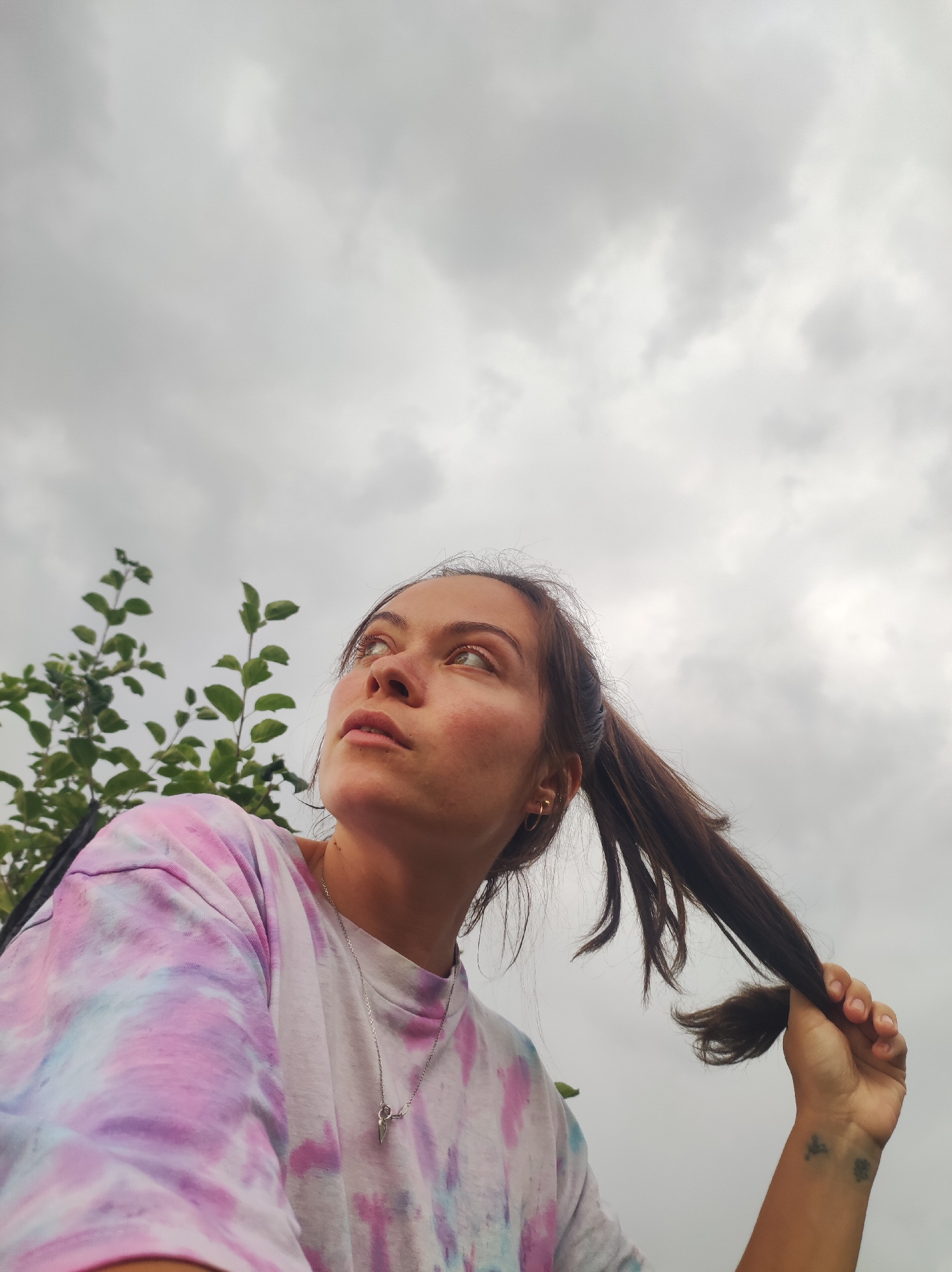 A young woman with brown hair pulled into a ponytail, wearing a tie-dye t-shirt, looks up at a cloudy sky. She is holding a section of her hair in her right hand. A green leafy plant is visible to her left. The image appears to be taken outdoors, possibly in or near the city of Fokino, Russia, given the provided context.