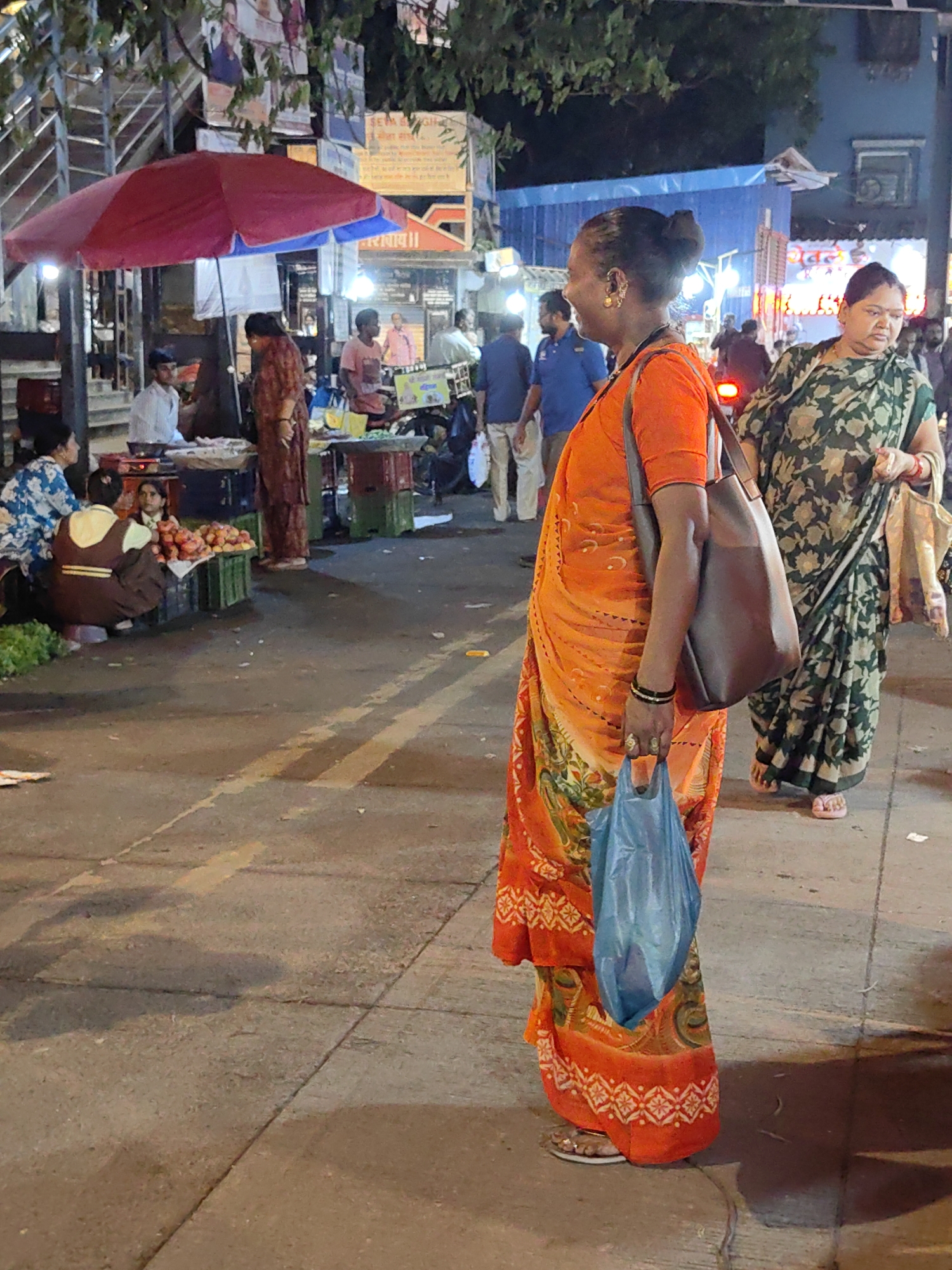 The image captures a street scene in Mumbai, India, likely in the evening given the artificial lighting and shadows. The foreground is dominated by two women dressed in traditional Indian attire. The woman on the right wears a dark green saree with a floral pattern and is holding a bag. She is facing left, looking slightly down. To her left, a woman in a vibrant orange saree with a patterned border is prominently featured in profile, facing left. She is carrying a dark brown shoulder bag and a light blue plastic bag in her right hand. Her hair is tied up in a bun. In the background, various stalls are visible, suggesting a market or street food area. A red and blue umbrella shades one of the stalls where a man in a white shirt appears to be attending to customers. Another person in a maroon outfit is standing near the stall. Further in the background, more people and stalls are dimly lit, with signs and posters adorning the overhead structures. The ground is paved, with yellow lines indicating pedestrian paths or traffic dividers. The overall atmosphere is that of a bustling urban market at dusk.