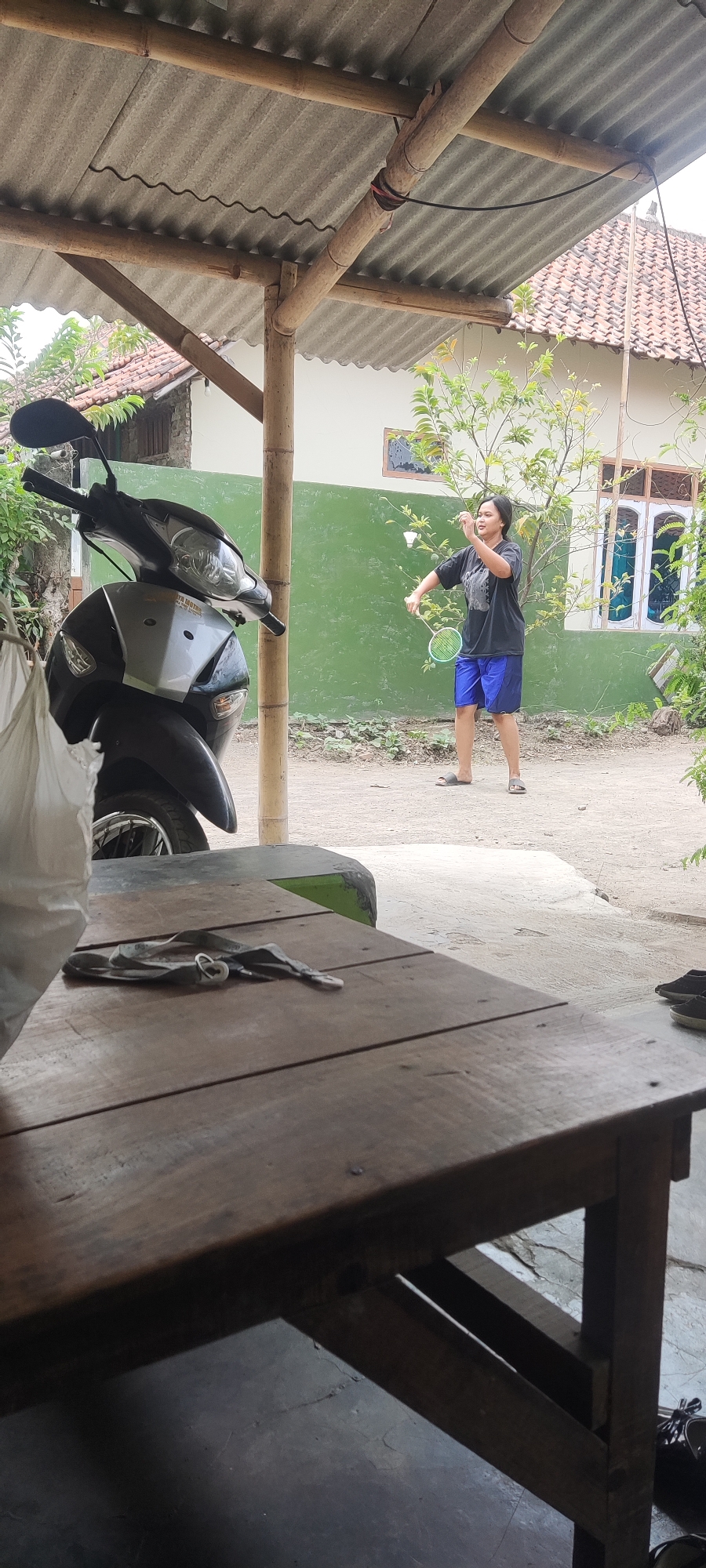 In Karangwangun, Indonesia, a young woman is captured in a moment of playing badminton. She stands in a yard, dressed in a black t-shirt and blue shorts, holding a badminton racket and positioned as if to strike a shuttlecock. The environment includes a residential house with green walls and a tiled roof, a scooter parked to the left, and a wooden table in the foreground. The scene suggests a casual, outdoor activity within a residential setting.