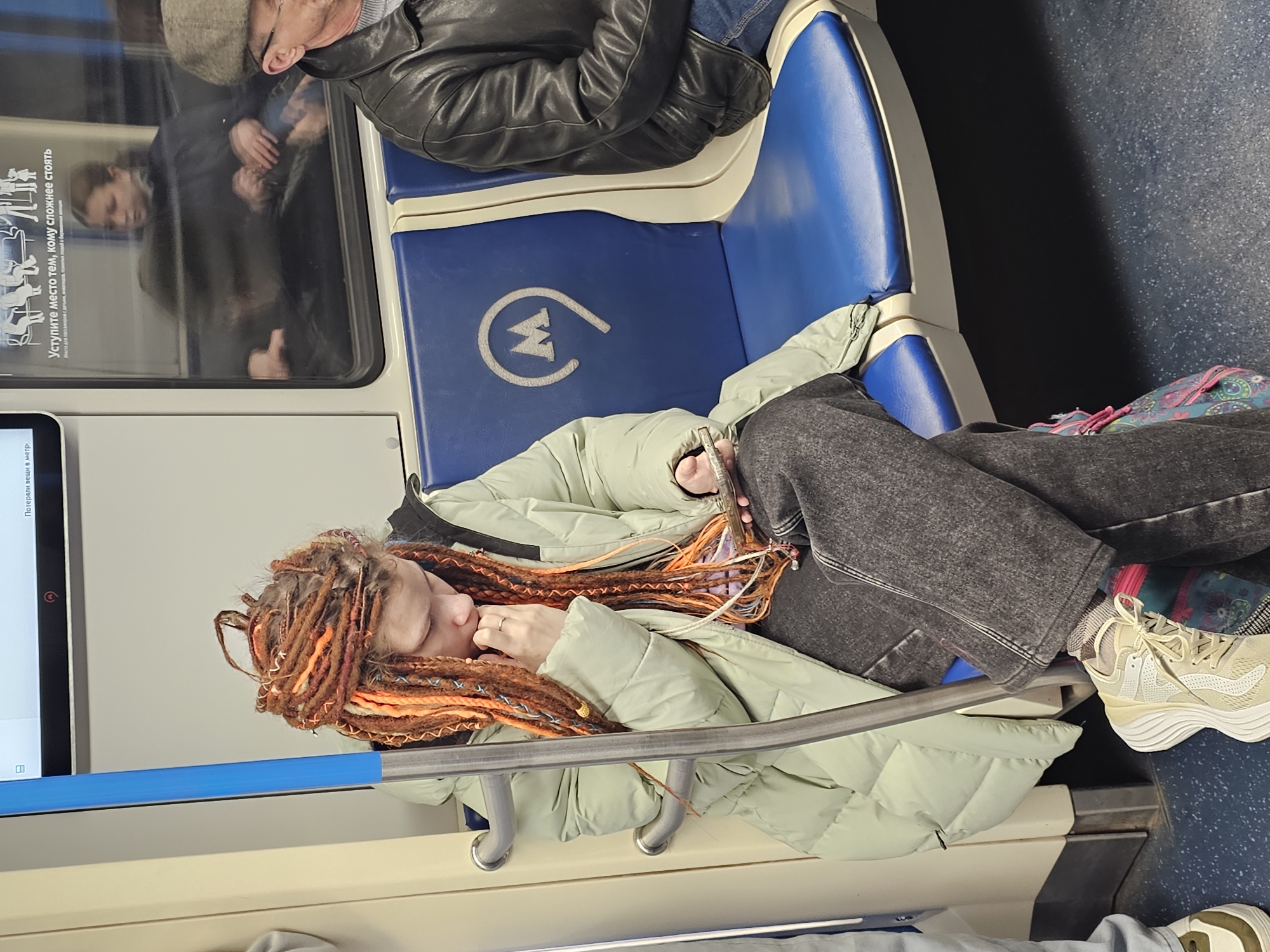 This image captures a scene inside a Moscow Metro subway car. In the foreground, a young person with long, distinctive reddish-orange and brown dreadlocks is seen. They are wearing a light green puffy jacket, dark gray wide-leg pants, and light-colored athletic shoes, and are engrossed in looking at a smartphone while resting on a blue subway seat. A colorful, patterned backpack or bag is visible on the floor next to them.
Behind the main subject, an older man wearing a flat cap and a dark leather jacket is seated on an adjacent blue seat. The blue seats, with their cream-colored trim, prominently display the "M" logo, which is characteristic of the Moscow Metro.
To the left, through the subway car window, reflections of other passengers are visible, including a person in a dark coat and a hand making a thumbs-up gesture. A white sign affixed to the window contains Cyrillic text, reading "Уступите место тем, кому сложнее стоять" (Yield your seat to those for whom it is difficult to stand) and "Места для пассажиров с детьми," (Seats for passengers with children,). A digital display on the lower left side shows the text "Потеряли вещи в метро" (Lost items in the metro). The interior features standard metal handrails and a speckled dark floor, indicating a public transport setting during what appears to be daytime.