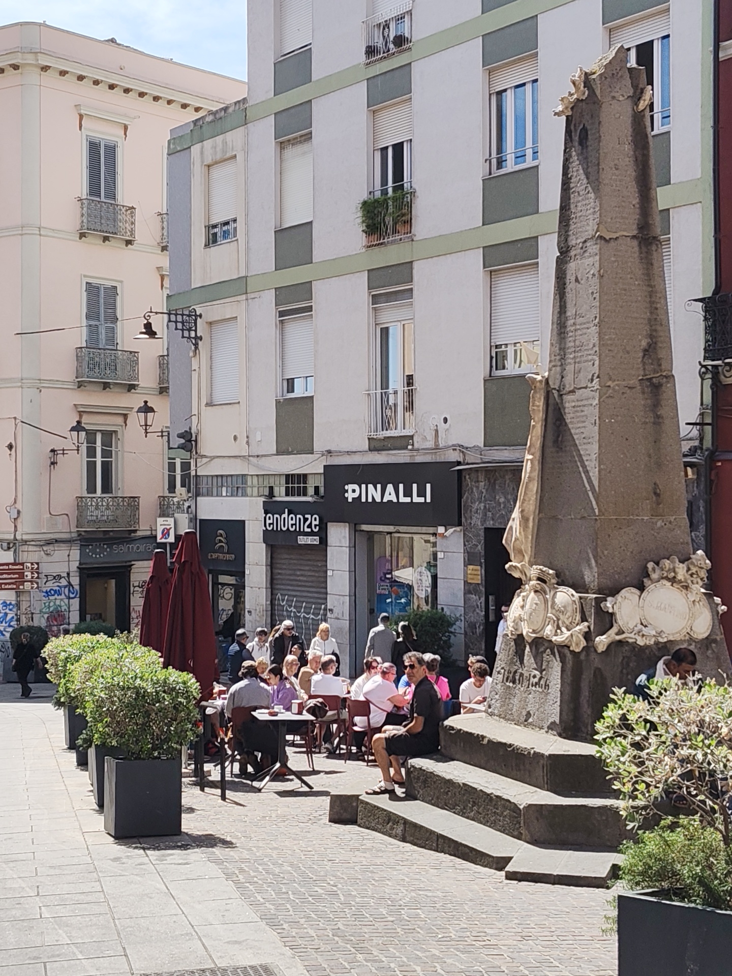 The image captures a street scene in Cagliari, Italy. In the foreground, there's a weathered stone monument with ornamental carvings and what appears to be inscriptions. A man is seated on the monument's steps. Several people are seated at outdoor tables, enjoying a meal or drinks, under the shade of large red umbrellas. The dining area is defined by potted green bushes.
Behind the dining area, a storefront labeled "PINALLI" is visible. The buildings in the background have a classic European architectural style, featuring multiple stories and windows. The street is paved and appears to be a pedestrian zone. The weather seems sunny and bright, with clear skies.