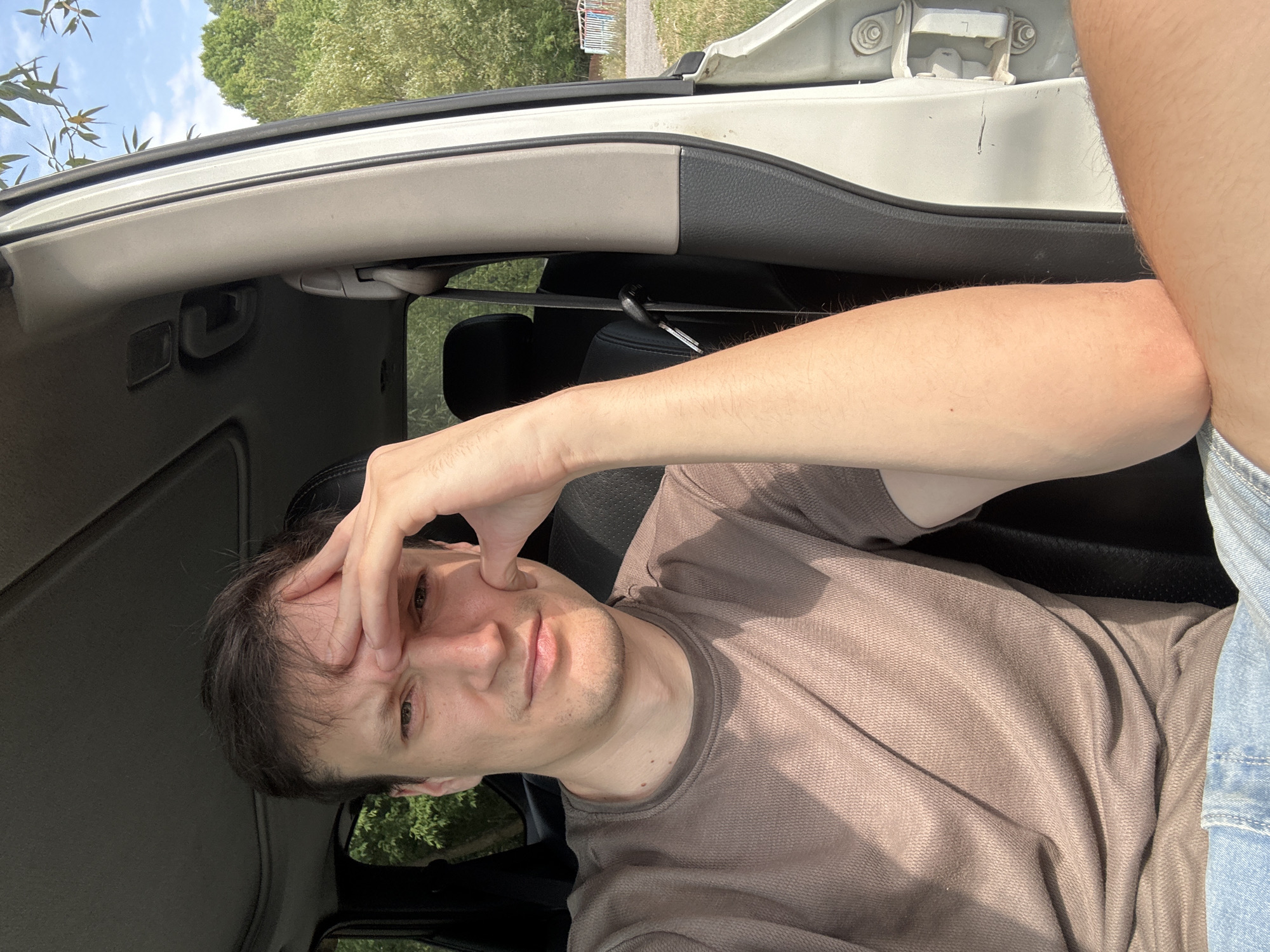 A man sits in the driver's seat of a vehicle, with the car door open. He is wearing a brown t-shirt and denim shorts, and his left hand is placed on his forehead, suggesting he might be tired or stressed. The background shows trees and a bright, sunny sky, indicating an outdoor setting. It is not possible to determine if the location is the city of Makiv, Ukraine, from the provided image.