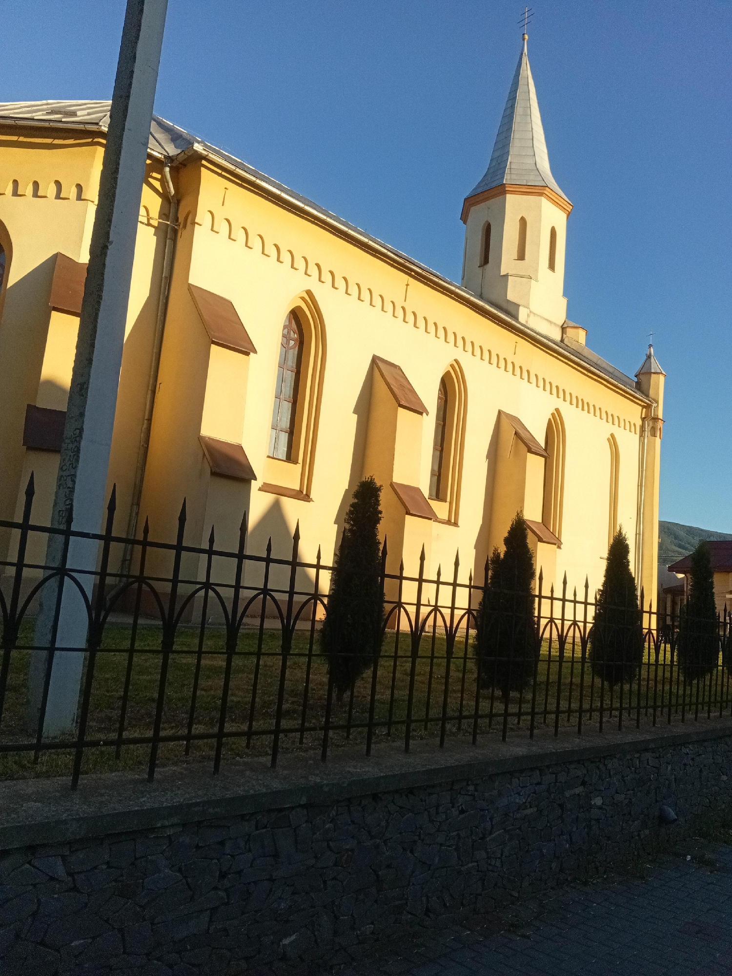 The image shows a yellow church with a tall, pointed steeple against a clear blue sky. The church has arched windows and decorative trim along the roofline. A black metal fence runs in front of the church, with several conical evergreen trees visible behind it. A concrete utility pole is positioned in the foreground, partially obscuring the left side of the church. The church is located in Tiachiv, Ukraine. The sun is casting long shadows, suggesting it is either early morning or late afternoon. The ground behind the fence appears to be a lawn, and a stone wall runs along the bottom of the image.