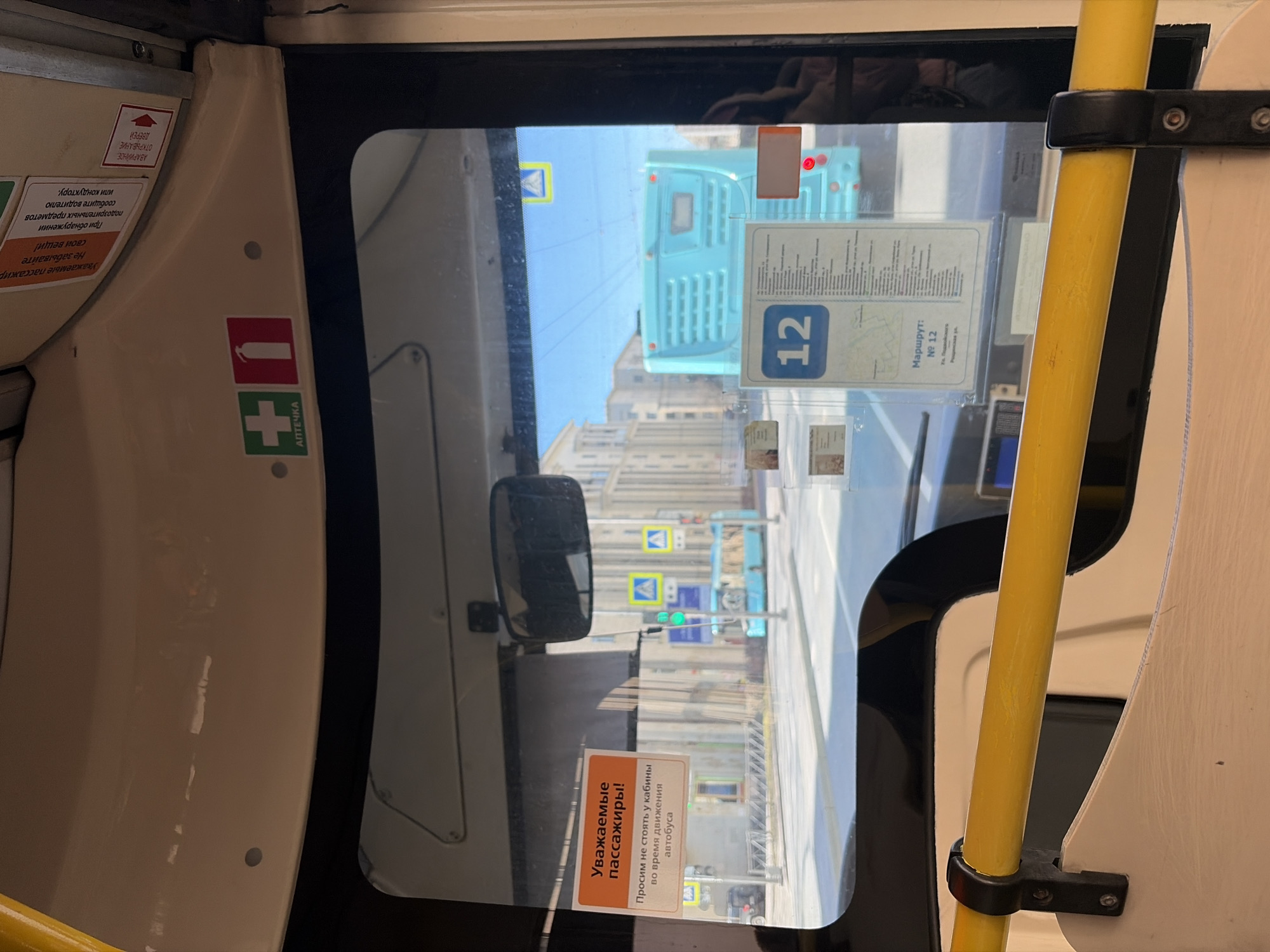 The image shows a view from inside a bus in Saint Petersburg, Russia. The outside view is visible through the window, depicting a street scene with buildings, traffic lights, and a turquoise bus ahead. Inside the bus, several details are visible, including stickers with Russian text ("Уважаемые пассажиры! Просим не стоять у кабины во время движения автобуса" - "Dear passengers! Please do not stand by the driver's cabin while the bus is moving") and route information ("Маршрут: № 12" - "Route: No. 12"). The "Аптечка" ("First Aid Kit") sticker is visible. There is a yellow handrail mounted next to the window.
