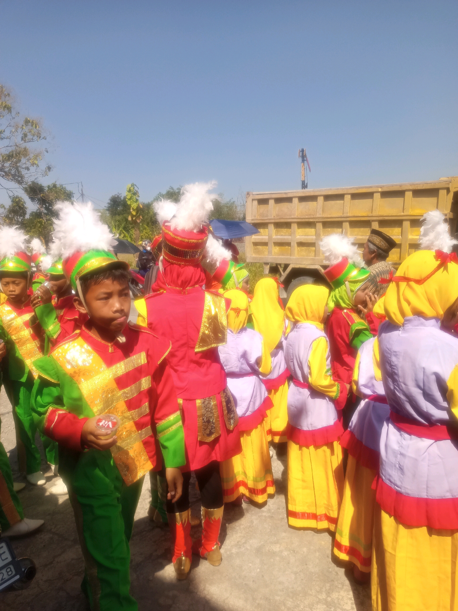 A group of children dressed in brightly colored marching band uniforms stand outdoors under a clear blue sky. The uniforms consist of red jackets with gold trim, green pants, and green and red hats adorned with white feathers. Some of the children are holding small, round objects. In the background, a large yellow dump truck is parked, and there are trees and other vegetation visible. The ground appears to be paved with concrete or asphalt. The image seems to capture a moment before or after a parade or performance, given the formal attire and the gathering of participants. The location is specified as Sumberlawang, Indonesia.