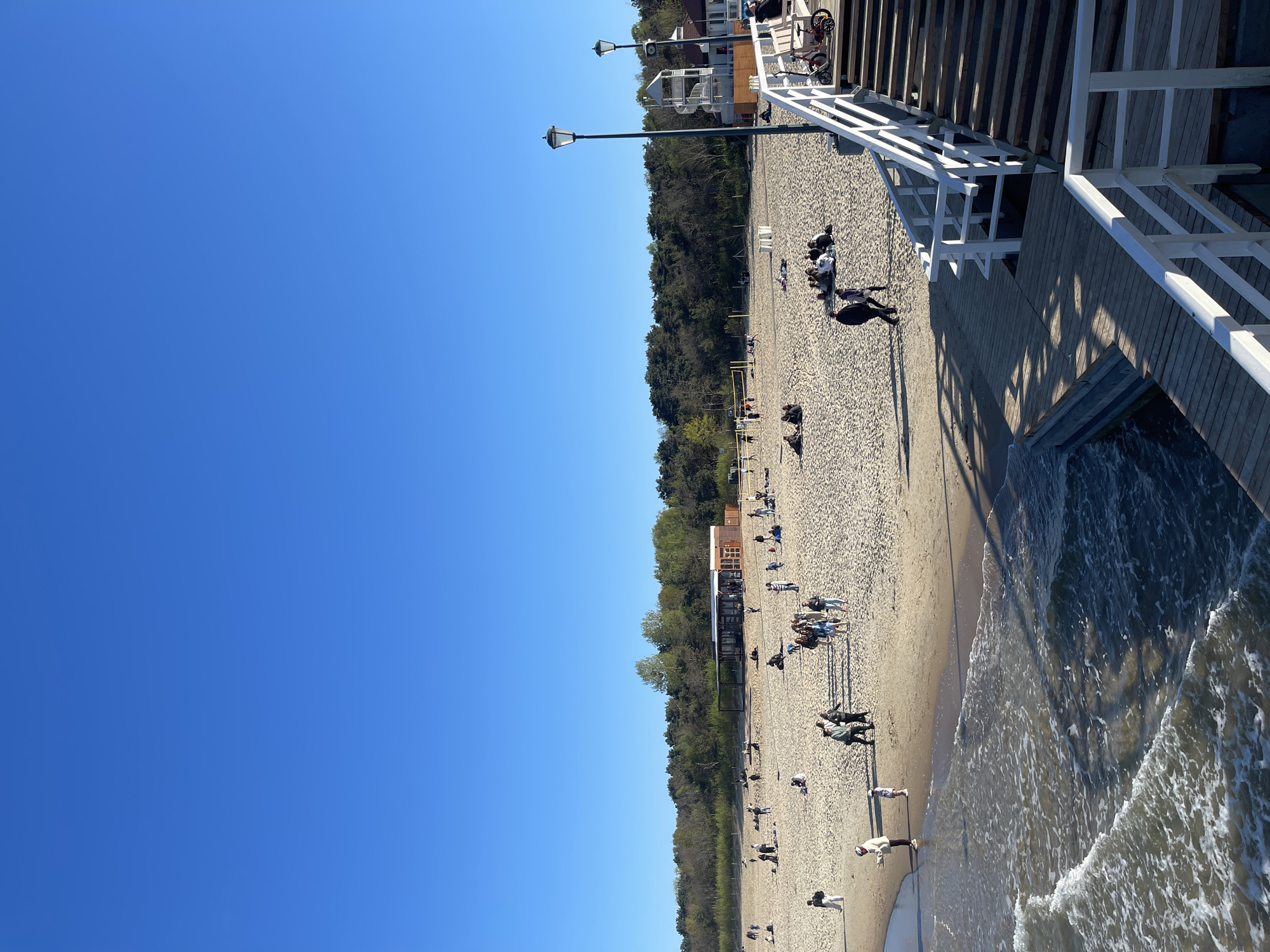 This image shows a beach scene in Gdańsk, Poland. The beach is crowded with people. Some are sunbathing or resting. Others are walking, and some are near the water. The shoreline is visible, with gentle waves coming to shore. A forested area runs along the back of the beach. A wooden pier extends from the right side of the image, with white railings and stairs leading down to the sand. The sky is clear and blue. The image appears to be taken from an elevated position on the pier.