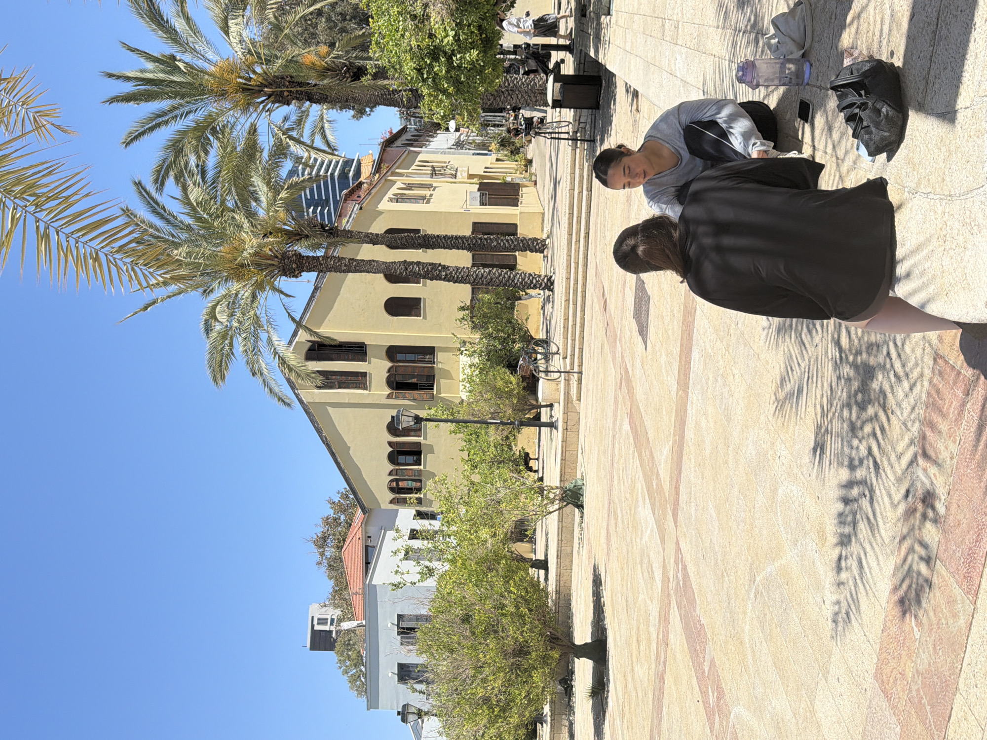 The image shows a street scene in Tel-Aviv, Israel, characterized by Mediterranean architecture and palm trees. The sky is clear and blue.
Two women are on the right side of the image, seated on a stone-paved walkway. One woman is wearing a light gray long sleeve, dark athletic pants and has her hair pulled back and appears to be looking at something on the ground. The other woman is wearing a black top and shorts. Her hair is pulled back and is walking away from the seated woman.
A backpack, water bottle and a cell phone are placed next to the woman in gray.
Behind the women, there is a row of buildings, primarily yellowish in color with characteristic arched windows. The buildings are three stories tall. Palm trees are visible along the street. The stone-paved walkway has shadows of the palm trees.