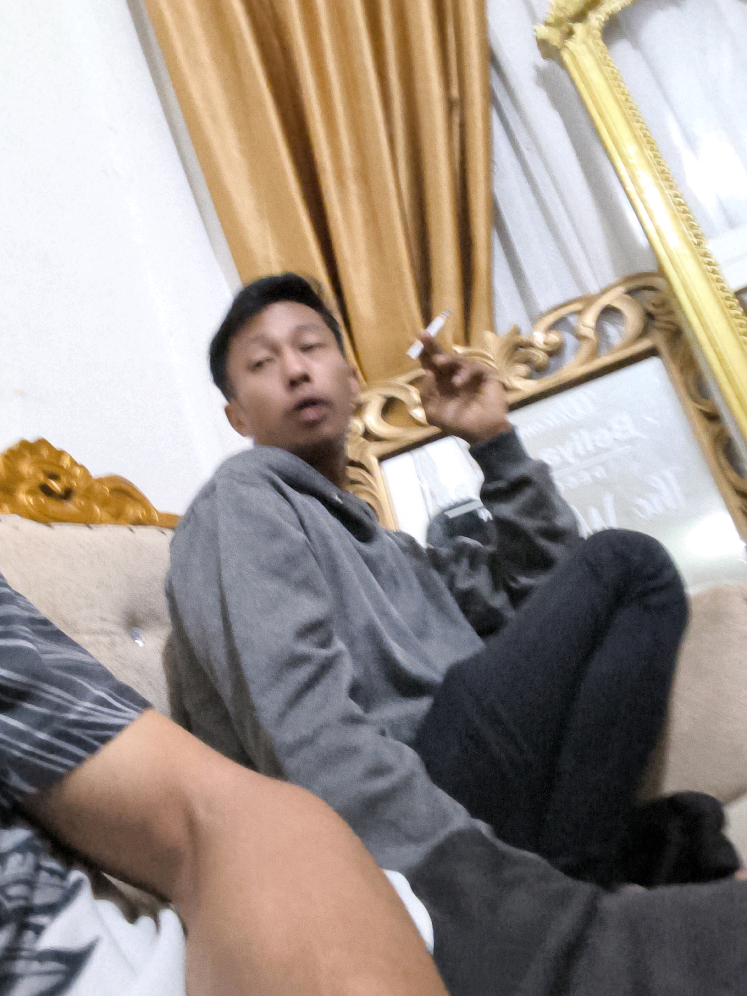 The image captures an indoor scene featuring two individuals. One person, a young man with short dark hair, is seated on a sofa and holding a cigarette in his right hand, which is raised to his mouth. He is wearing a grey hooded sweatshirt and dark pants. The other person's arm is visible in the foreground, partially obscuring the view of the first individual's legs.
The background reveals a luxurious gold-framed mirror and draped golden curtains. The sofa appears to be upholstered in a light-colored fabric. The overall ambiance suggests a casual setting within a home. The location, Koto Baru Semurup, Indonesia, provides the geographical context for this scene.