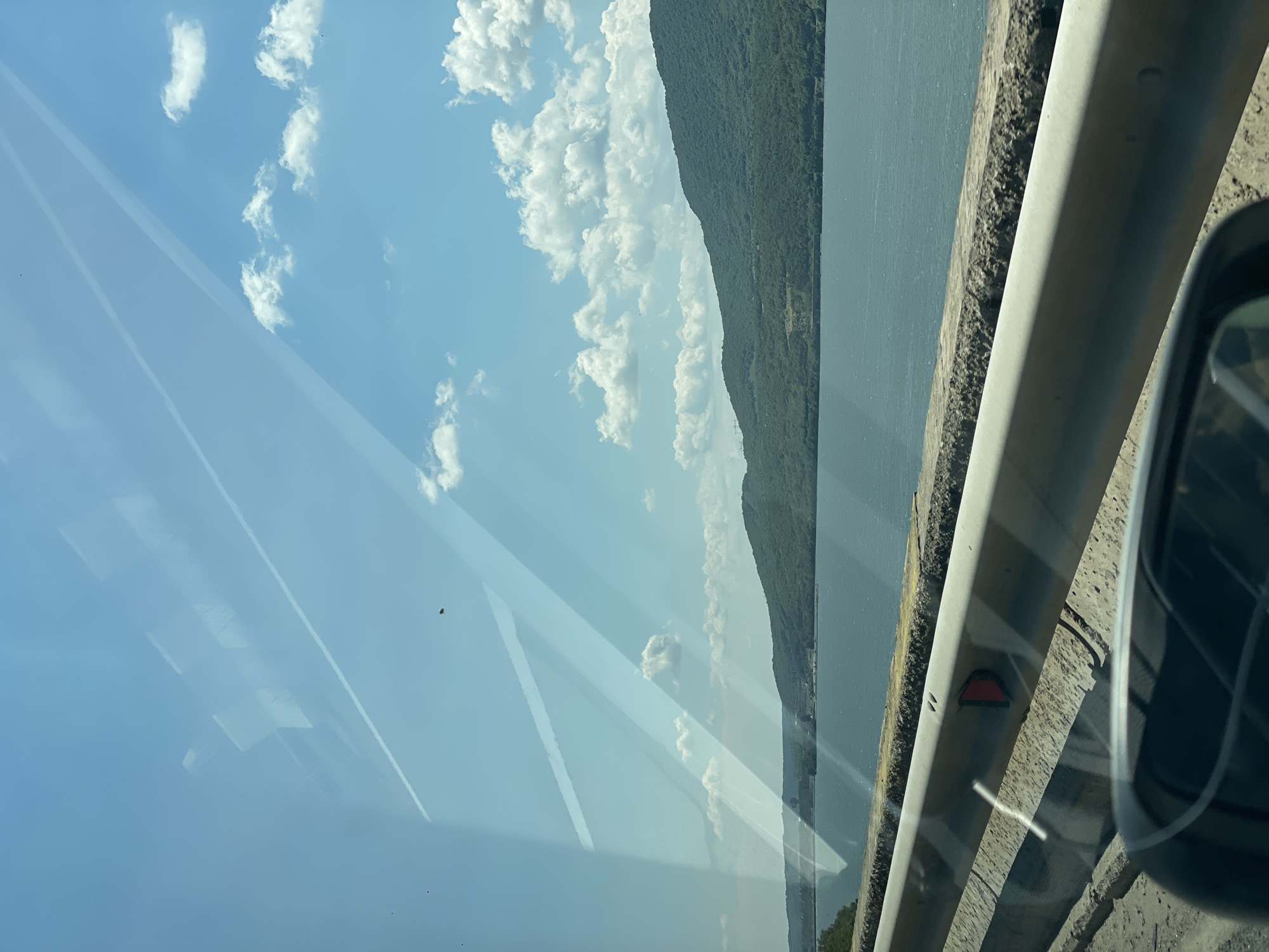 The image depicts a landscape viewed from the interior of a vehicle, positioned alongside a large body of water with forested hills beyond, near Drobeta-Turnu Severin, Romania. The clear blue sky, visible in the upper two-thirds of the frame, is dotted with scattered white cumulus clouds. Two small, dark, irregular specks are present against the sky, one in the upper center and another lower and to the right. The vehicle's windshield exhibits prominent diagonal light reflections and faint geometric reflections, partially obscuring the view. A subtle, thin white line, possibly a contrail reflection, is visible in the upper left.
The middle ground features a wide expanse of dark blue-green water, consistent with the Danube River in the specified location. On the far side of the water, densely forested green hills or mountains rise, extending across the entire background. Faint structures, potentially industrial or related to power generation (consistent with the Iron Gates I Hydroelectric Power Station nearby), are discernible on the distant left bank.
In the foreground, a light gray metallic guardrail runs horizontally across the lower portion of the image, atop a textured, weathered concrete barrier. A red, triangular reflector is affixed to the guardrail, pointing to the left. The reflection of a dark car side mirror is visible in the bottom right corner, revealing a partial view of the road surface and the vehicle's interior. The ground immediately adjacent to the guardrail appears to be rough concrete or asphalt.