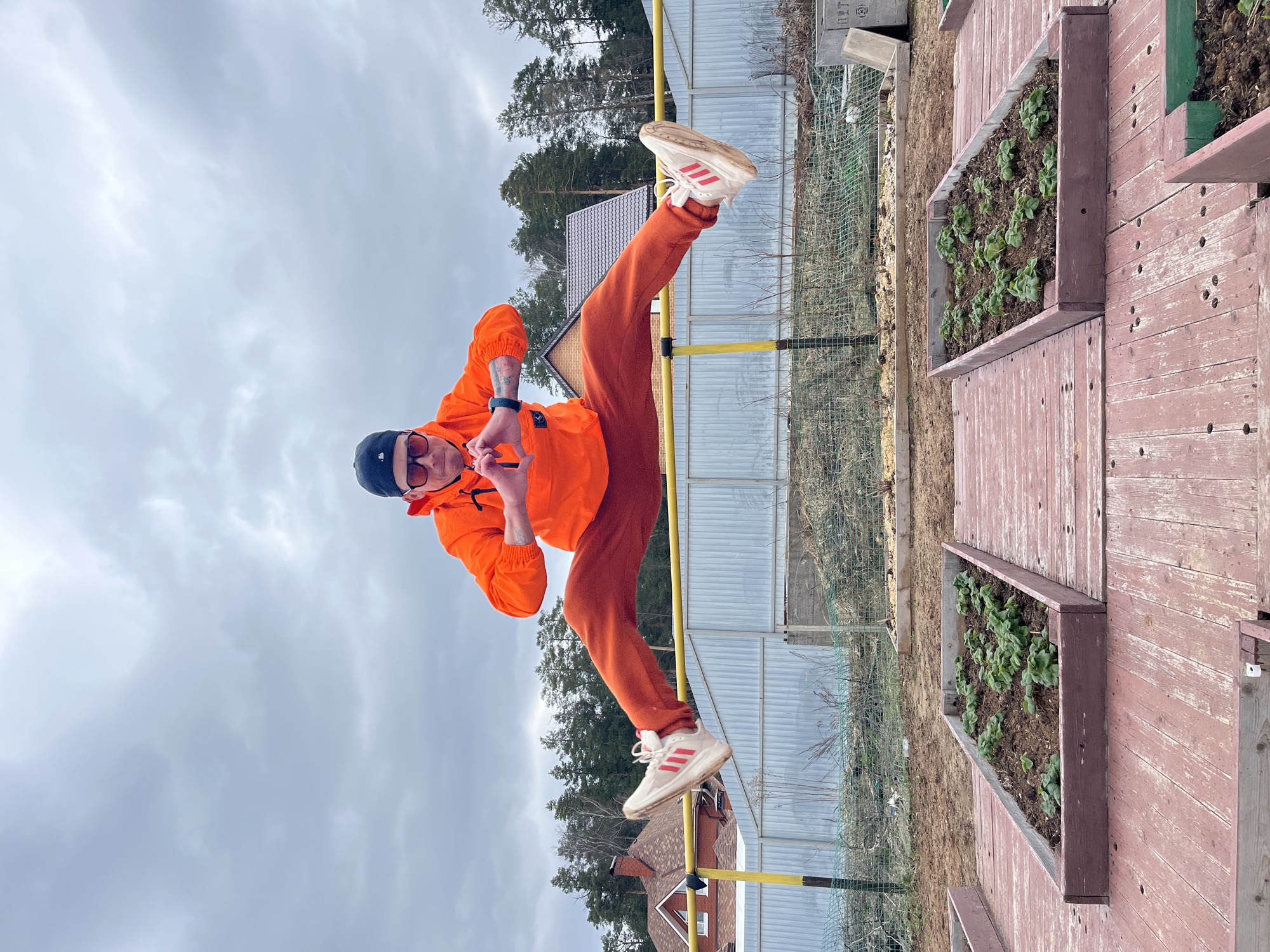 A man in an orange tracksuit, a black beanie, and sunglasses is balancing precariously on a yellow metal bar, with one leg extended high. The setting appears to be a residential area, possibly a garden, near Chaikovsky, Russia. Visible in the background are raised garden beds containing plants, a wire fence, trees, houses, and a cloudy sky.