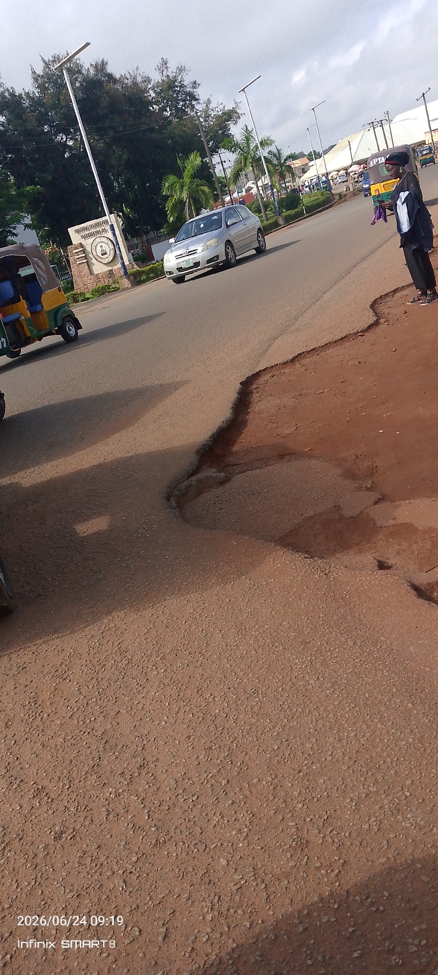 In the city of Jos, Nigeria, on June 24, 2026, at 09:19 AM, the scene depicts a street with a large pothole in the foreground, indicative of poor road conditions. To the left, a three-wheeled auto-rickshaw, commonly known as a Keke Napep, is visible. Further down the road, a silver sedan is driving past a sign that reads "UNIVERSITY OF JOS". Across the street, a person in dark clothing stands on the sidewalk, holding a purple object. Palm trees and other vegetation line the side of the road, and streetlights are present. The sky is overcast with some clouds. The overall environment suggests an urban setting with moderate traffic.