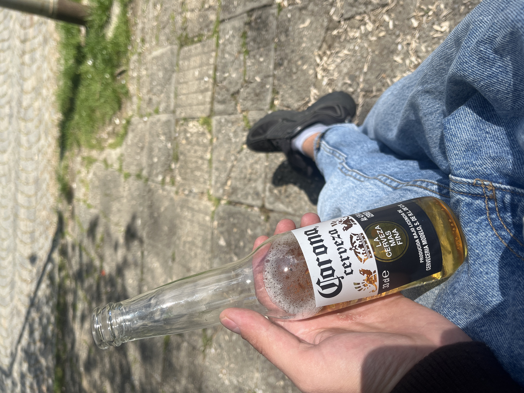 This angled close-up image shows a person outdoors in Sofia, Bulgaria, holding a partially consumed Corona beer bottle. The individual's lower body is visible, featuring blue denim jeans and black shoes with white socks. Their hand cradles the clear glass bottle, which contains a small amount of golden liquid and a layer of foam at the bottom.
The bottle's label is prominent, displaying "Corona cerveza," "LA CERVEZA MÁS FINA," "Alc 4.5%," "33 cl," and "PRODUCIDA BAJO LICENCIA DE CERVECERIA MODELO. S. DE R.L. DE C.V."
The setting appears to be a paved outdoor area, possibly a sidewalk or path, made of light-colored stone or concrete pavers. Green grass and dirt are visible between the paving stones. The scene is brightly illuminated by natural sunlight, indicating a clear and sunny day, likely mid-morning or afternoon. A blurry, dark metallic object, potentially a pipe or railing, is present in the upper left background. The overall mood suggests a casual moment of relaxation.