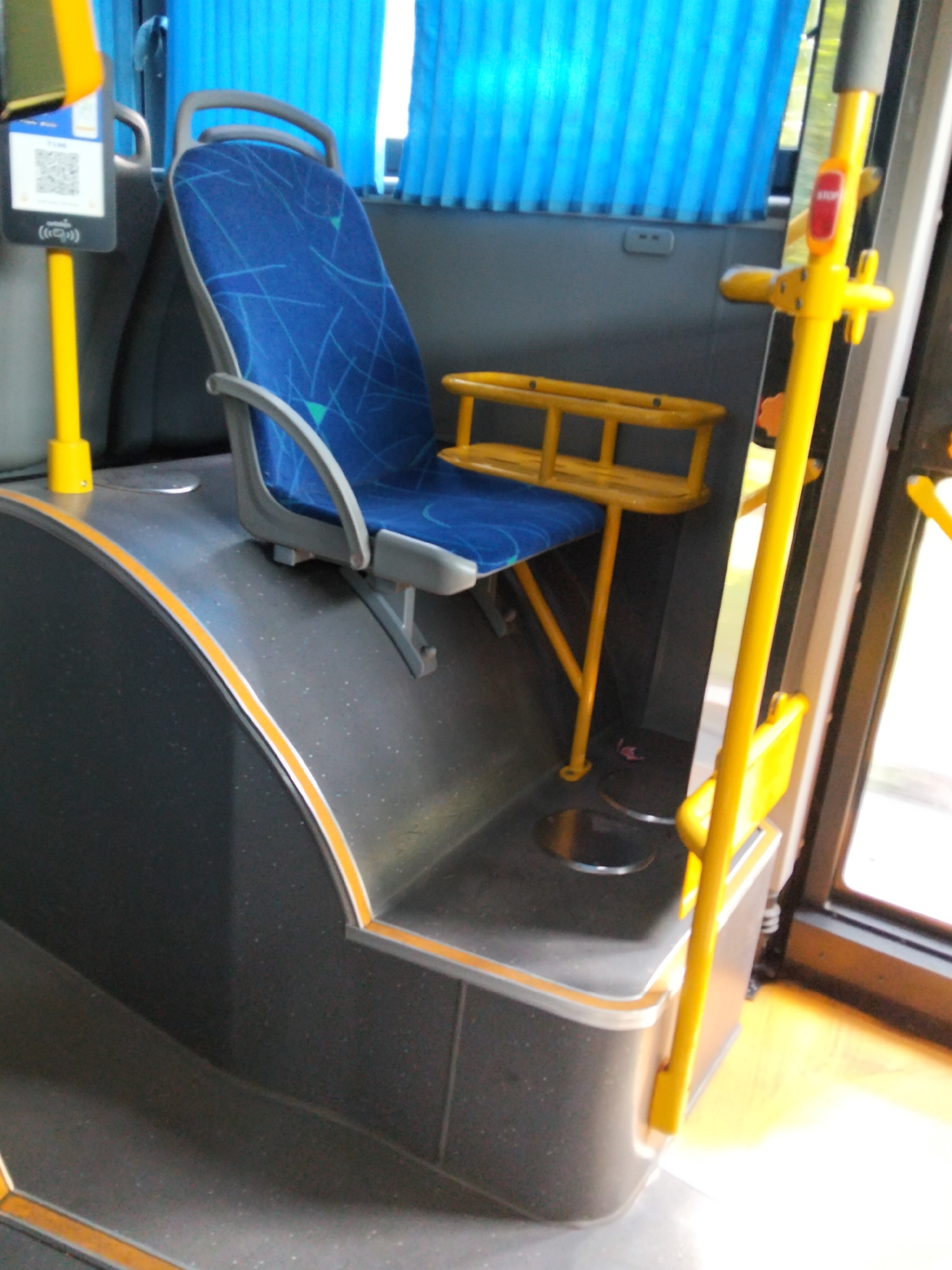 The image shows the interior of a public transport vehicle, likely a bus, in Almaty, Kazakhstan. The focal point is a blue fabric seat with a patterned design, positioned on a grey, textured floor. Adjacent to the seat, a yellow metal pole rises vertically, equipped with a red stop button. Another yellow horizontal bar is attached to the vertical pole, providing a handrail. A small yellow rack, possibly for luggage or personal items, is mounted near the seat. The background features blue curtains and a glimpse of the exterior through a window. The overall impression is of a functional and clean interior space within a public transportation system.