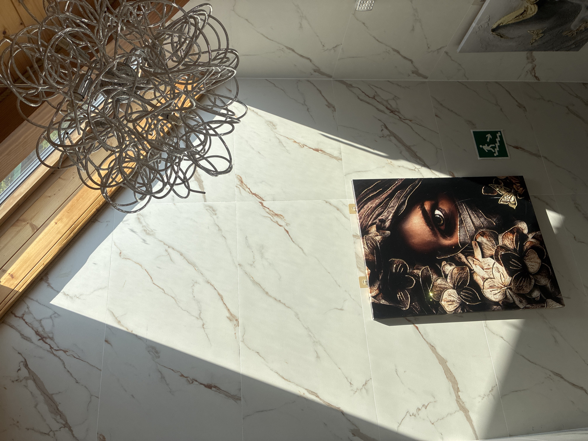 The image shows an interior scene with marble-patterned walls. A large, abstract, metallic light fixture hangs from the ceiling near a sloped wooden beam. A framed artwork depicting a woman's eyes peering from behind patterned fabric and flowers is mounted on the wall. Sunlight casts a strong diagonal shadow across the wall and artwork. A green exit sign with a running figure and arrows is also visible on the wall. The location is identified as Estosadok, Russia.