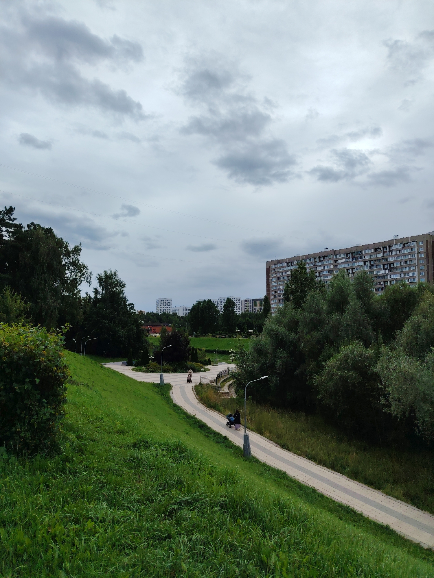 The image depicts a park scene in Zelenograd, Russia, under a cloudy sky. In the foreground, a grassy slope curves to the left. A paved pathway runs along the middle ground, bordered by a darker green area and trees on the right. On the pathway, a person pushes a stroller. Further back, on the pathway, another person walks, and in the distance, several multi-story apartment buildings are visible, characteristic of the area. The overall atmosphere is overcast.