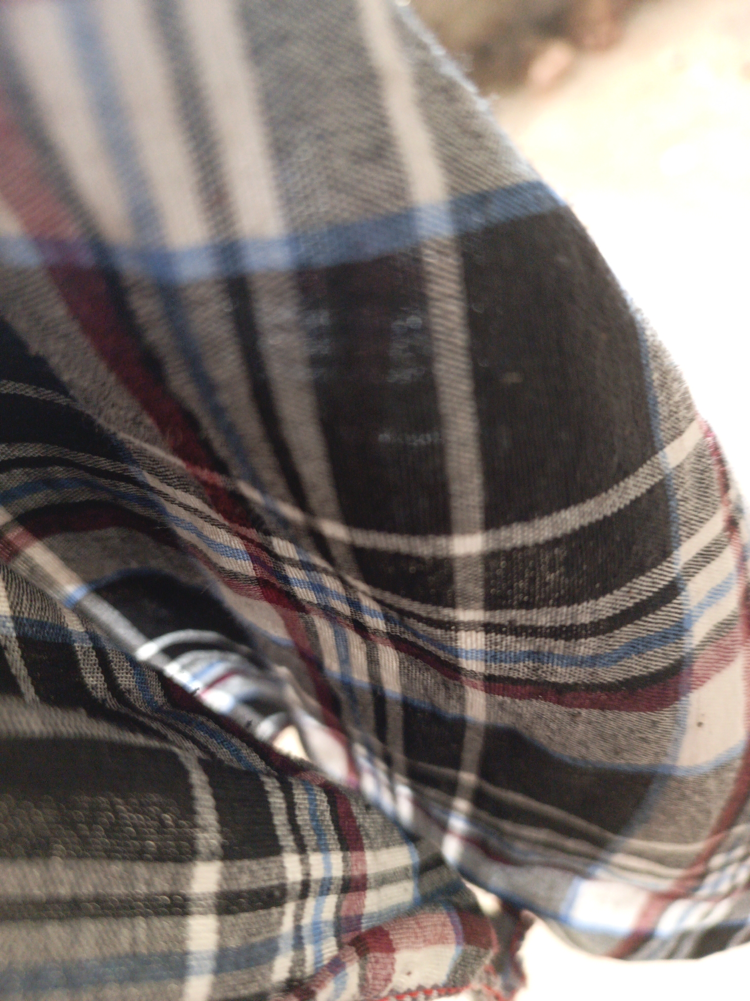 This close-up image features a plaid fabric, likely from an article of clothing or a blanket, draped in soft folds and creases. The pattern consists of stripes in varying widths and shades, including black, white, gray, blue, and burgundy or deep red. The fabric appears to have a slightly textured surface, possibly indicating material like flannel or cotton.
The foreground is sharply focused on the fabric, highlighting its woven pattern and the gentle undulations of its folds. The background is completely blurred and bright, suggesting a shallow depth of field and a well-lit environment, though no specific location cues are visible. There are no people or other distinct objects identifiable in the image, nor is any specific activity or event taking place. The image serves as a textural and pattern study of the fabric. No visible text is present.