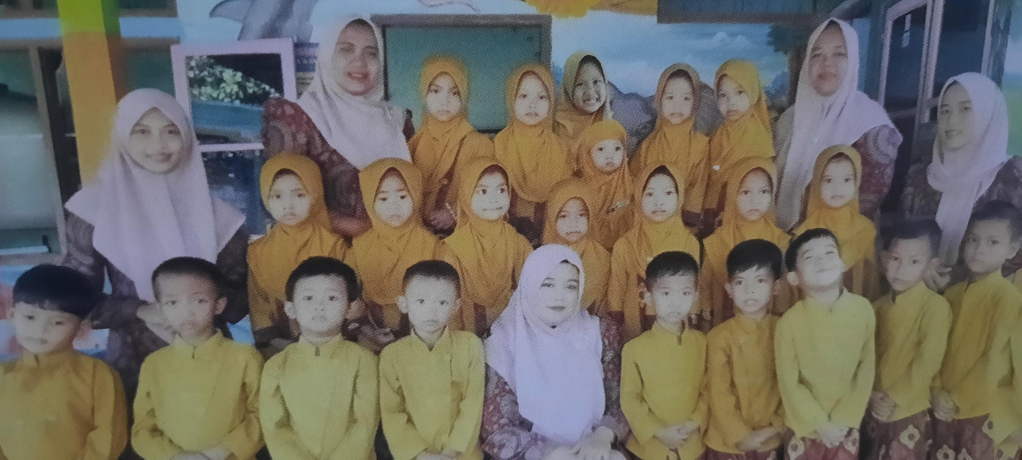 The image depicts a group of children and adults, likely posing for a photograph in what appears to be a school or community center setting. The location is identified as Ploso, Indonesia. The children are predominantly wearing yellow uniforms, with some girls also wearing hijabs in yellow and the adult women wearing hijabs in various colors, including white and light pink. Some of the children have dark hair, while others have lighter complexions. The background shows a colorful wall with what could be painted murals, possibly depicting nature scenes with a blue sky and mountains. There are also what appear to be framed pictures or artwork on the wall. The overall atmosphere suggests a formal gathering or an event, possibly a school picture day or a cultural celebration. The lighting is even, indicating it was likely taken indoors with artificial lighting or diffused natural light.