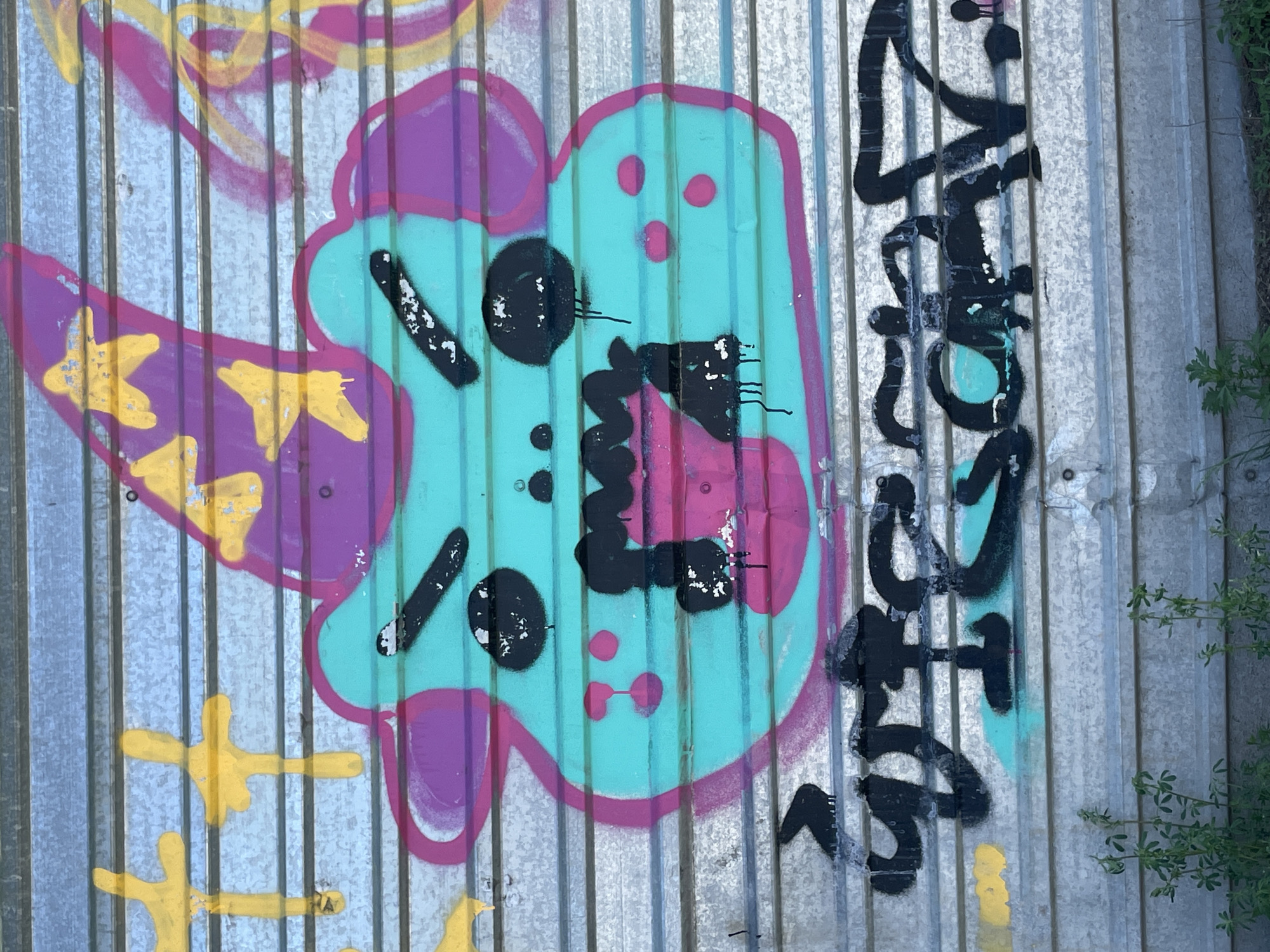 The image shows graffiti on a corrugated metal surface. The main figure is a stylized, cartoon-like face in teal, outlined in purple. It has black eyes and mouth, and pink accents. To the left are purple shapes with yellow markings. To the right is black graffiti lettering. The background is the metal sheeting. Some green foliage is visible on the right edge. The image does not contain sufficient information to determine the exact location within Izhevsk, Russia.