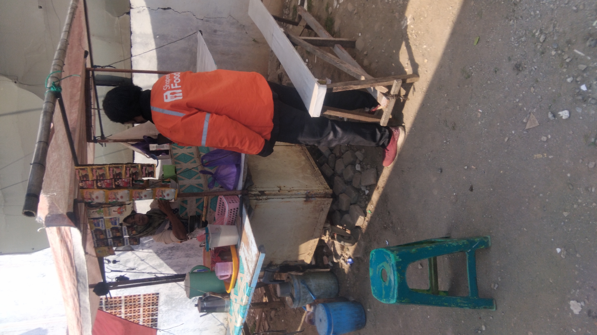 A street vendor is serving a customer at a stall in Batang, Indonesia. The vendor, a woman wearing a headscarf, stands behind a counter. In front of her are various items, including packaged snacks, drinks, and containers. A person wearing an orange jacket with the "Shopee Food" logo stands facing the vendor. The stall is covered by a tarp and is located on a dirt and gravel ground. A green plastic stool and a wooden bench are visible near the stall. The overall scene depicts a typical street-side food vending operation.