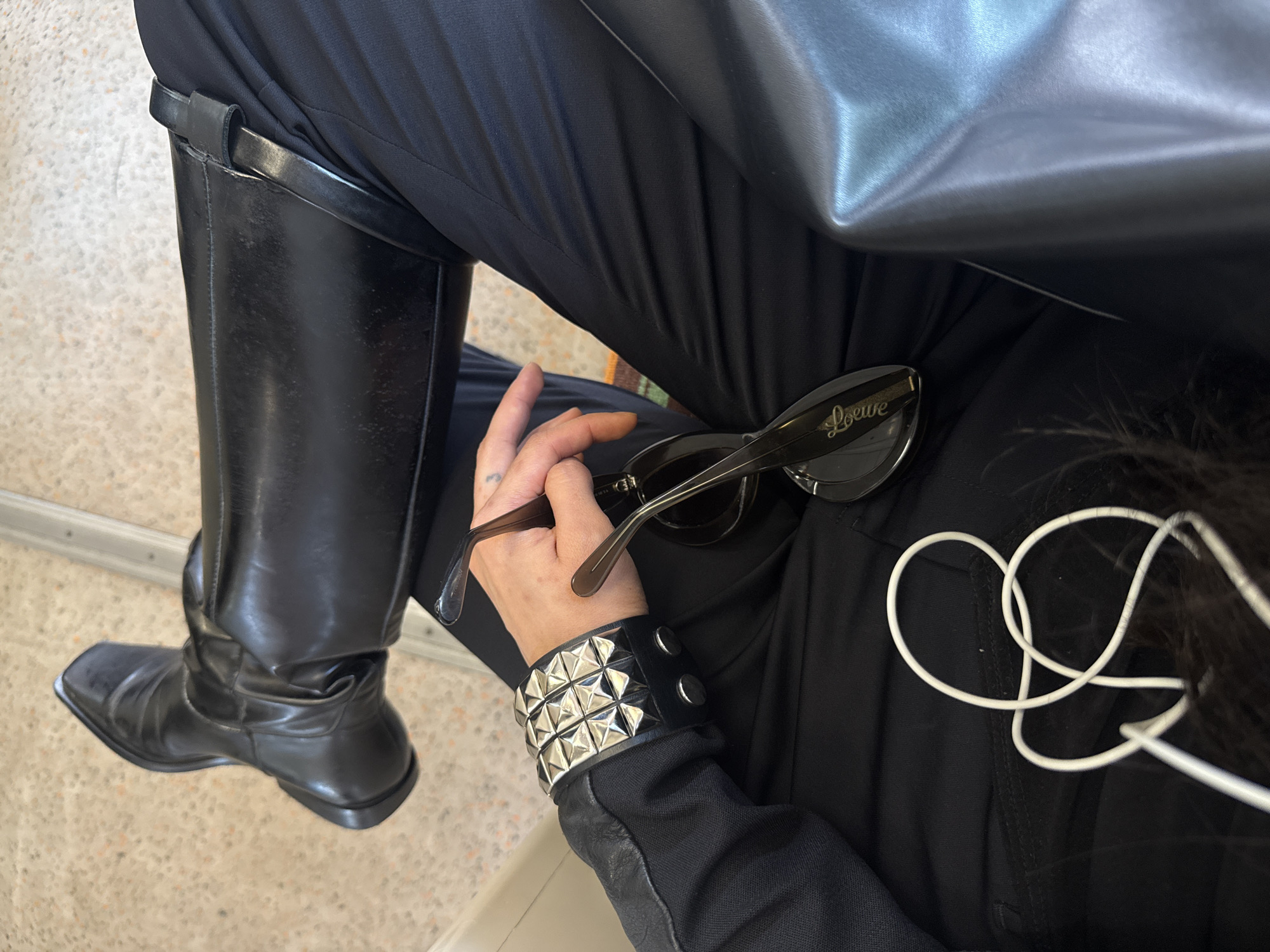 The image depicts a person seated, clad in black attire. They are wearing tall, black leather boots that reach to just below the knee. Their legs are covered in black pants. They are also wearing a black leather jacket. On their wrist, they have a black leather wristband studded with silver pyramids. In their hand, they are holding a pair of Loewe sunglasses with a transparent frame. White wired headphones are visible near their head. The location cannot be determined from the image.