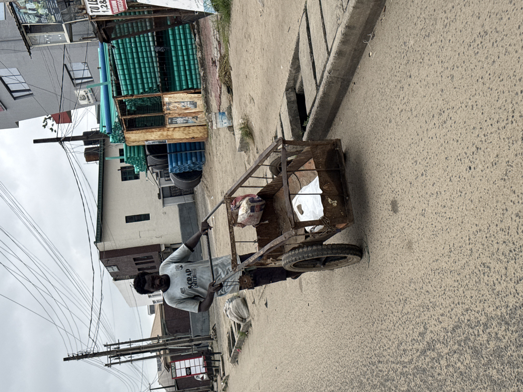 A man is pushing a rusty, two-wheeled cart loaded with items down a street in Ado, Nigeria. The man wears a light-colored t-shirt with graphics on it and dark shorts. The cart is made of metal and has a worn appearance. The street is paved and appears to be a busy area, with buildings and power lines visible in the background. Some of the buildings are modern, while others are older. There are also stacks of green pipes in the background, likely for construction or plumbing. The sky is overcast. The scene appears to be a typical urban street in Nigeria, with a mix of commercial and residential buildings.