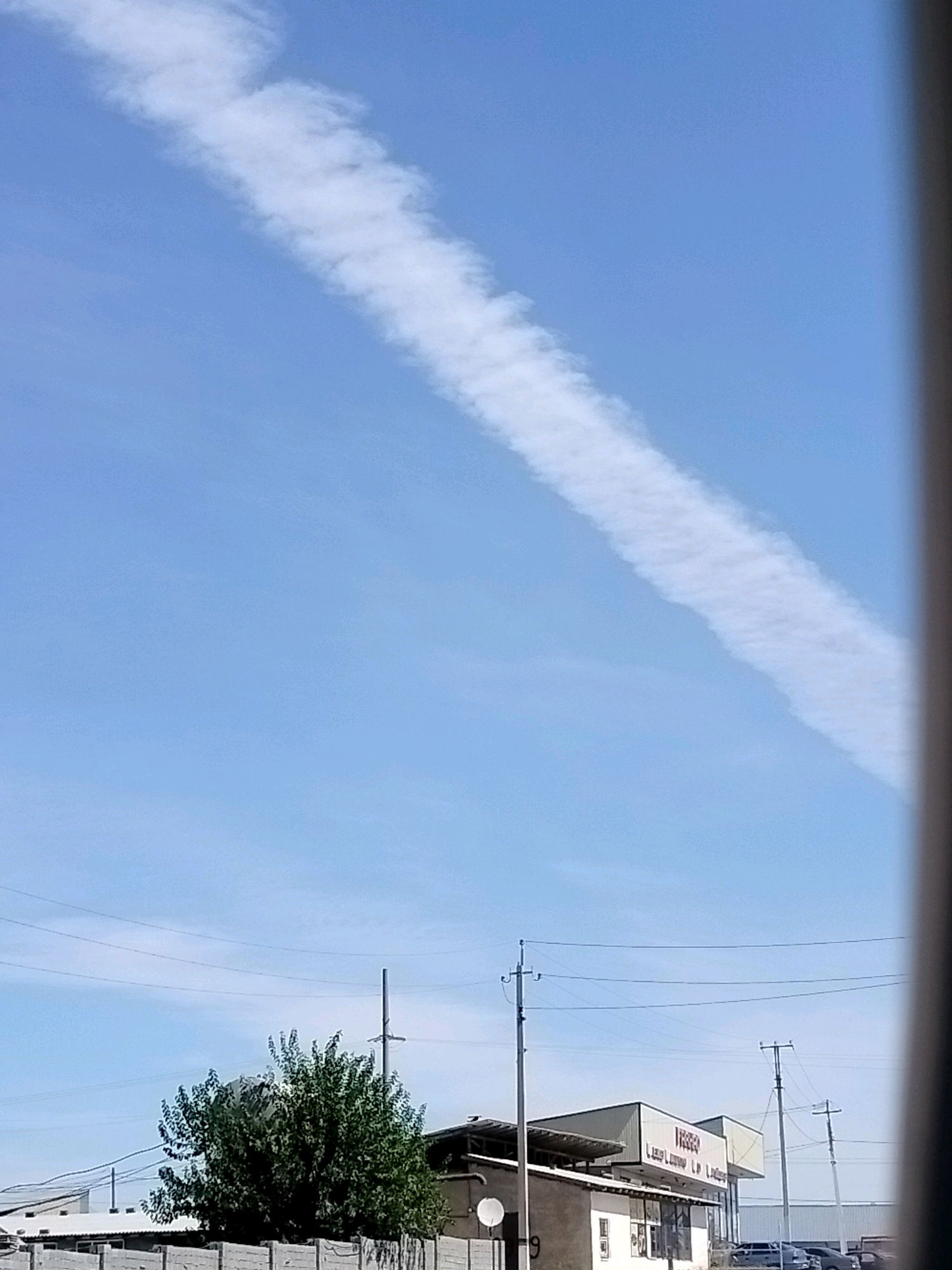 The image shows a clear blue sky with a long, thin white contrail stretching diagonally from the upper left to the lower right. In the lower portion of the image, there are several structures visible. A large, bushy green tree stands on the left, partially obscuring some buildings behind it. To the right of the tree, there is a building with a light-colored roof and a sign that reads "FOSTER" in red letters. Several utility poles with power lines are also present. The scene appears to be in an urban or industrial area. Given the context of Көлкент, Kazakhstan, this is likely a view of a street or an industrial zone within the city.