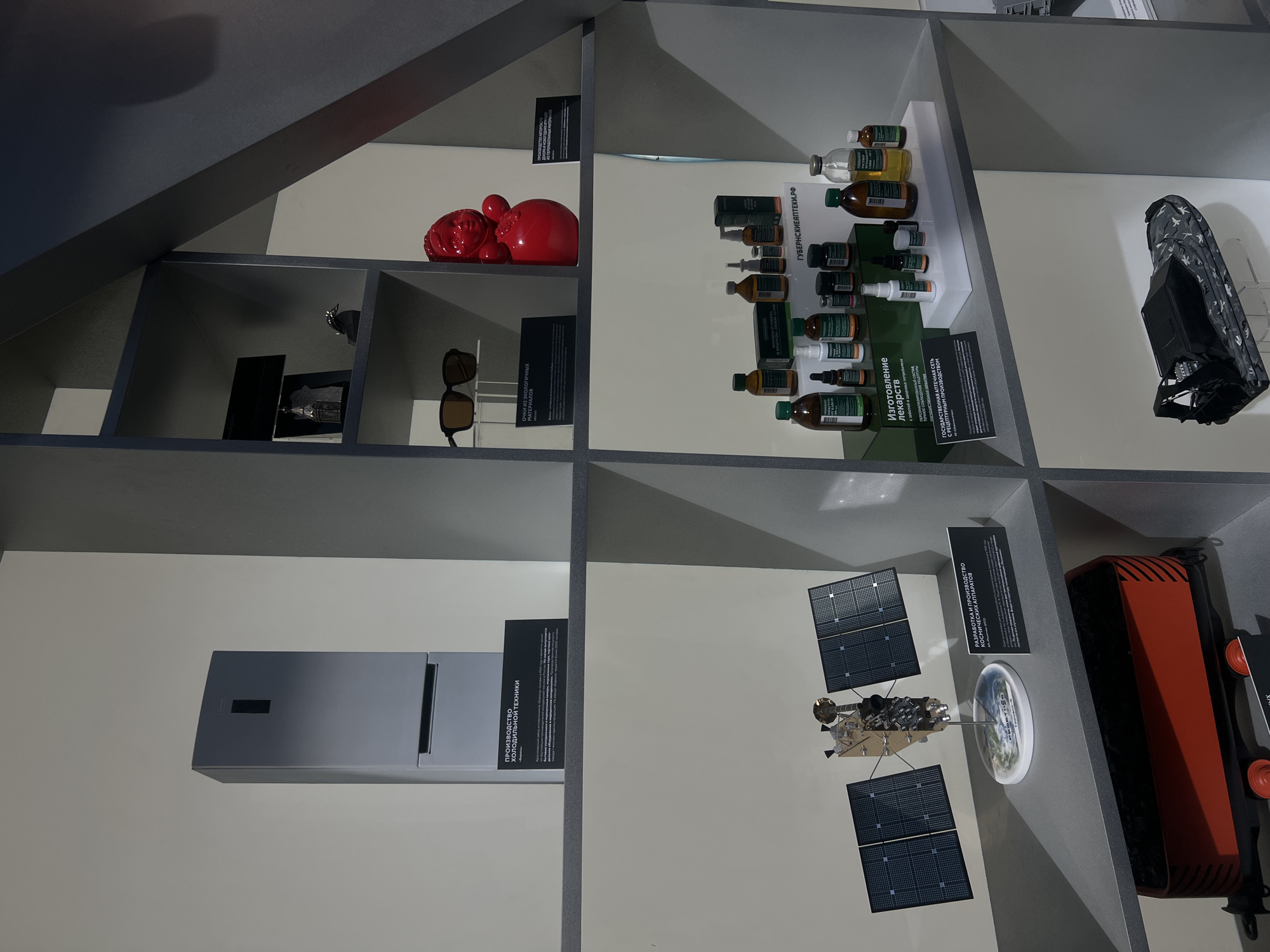 The image shows a display in what appears to be an exhibit, possibly a museum. The location is suggested by the signs in Russian, including the website "ГУБЕРНСКИЕАПТЕКИ.РФ," which indicates it might be in Russia, specifically Krasnoyarsk, as "Губернские аптеки" is a pharmacy chain in that city.
The display is constructed of grey shelving with various items on display.
Top Left Shelf: A red decorative object and next to it, a pair of sunglasses.
Middle Left Shelf: A grey rectangular object and next to it, a black sign with white Russian text mentioning "ПРОИЗВОДСТВО ХОЛОДИЛЬНОЙ ТЕХНИКИ" ("Production of Refrigeration Equipment").
Top Middle Shelf: A collection of bottled pharmaceuticals and next to it, a sign with Russian text mentioning "Изготовление лекарств с заботой о здоровье" ("Manufacturing of medicines with care for health").
Middle Middle Shelf: A satellite model, as well as a black sign with white Russian text mentioning "РАЗРАБОТКА И ПРОИЗВОДСТВО КОСМИЧЕСКИХ АППАРАТОВ" ("Development and production of spacecraft").
Top Right Shelf: A snowshoe.
Middle Right Shelf: A red apparatus, possibly a piece of equipment, with black stripes and orange wheels.
The overall impression is of a display showcasing local products, technologies, or history related to Krasnoyarsk.