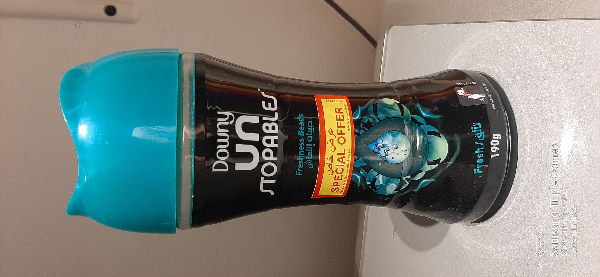 The image shows a bottle of Downy Unstopables fabric refresher beads with a special offer sticker. The bottle is dark and has a teal-colored cap. The label is black with white and teal text. A red and yellow sticker with the text "Special Offer" is prominently displayed. Below the sticker, there is an image of water droplets with blue and green swirls. The bottom of the bottle shows the text "Fresh / نَاقٍ" and "190g". A small symbol indicating that the product should not be thrown away with household waste is visible on the right side. The bottle is placed on a light-colored surface, possibly a shelf or countertop, with a shadow cast on it. The background is a plain, neutral color. The scene does not depict any recognizable landmarks or elements that would indicate a specific location within the city of Dubai, United Arab Emirates.