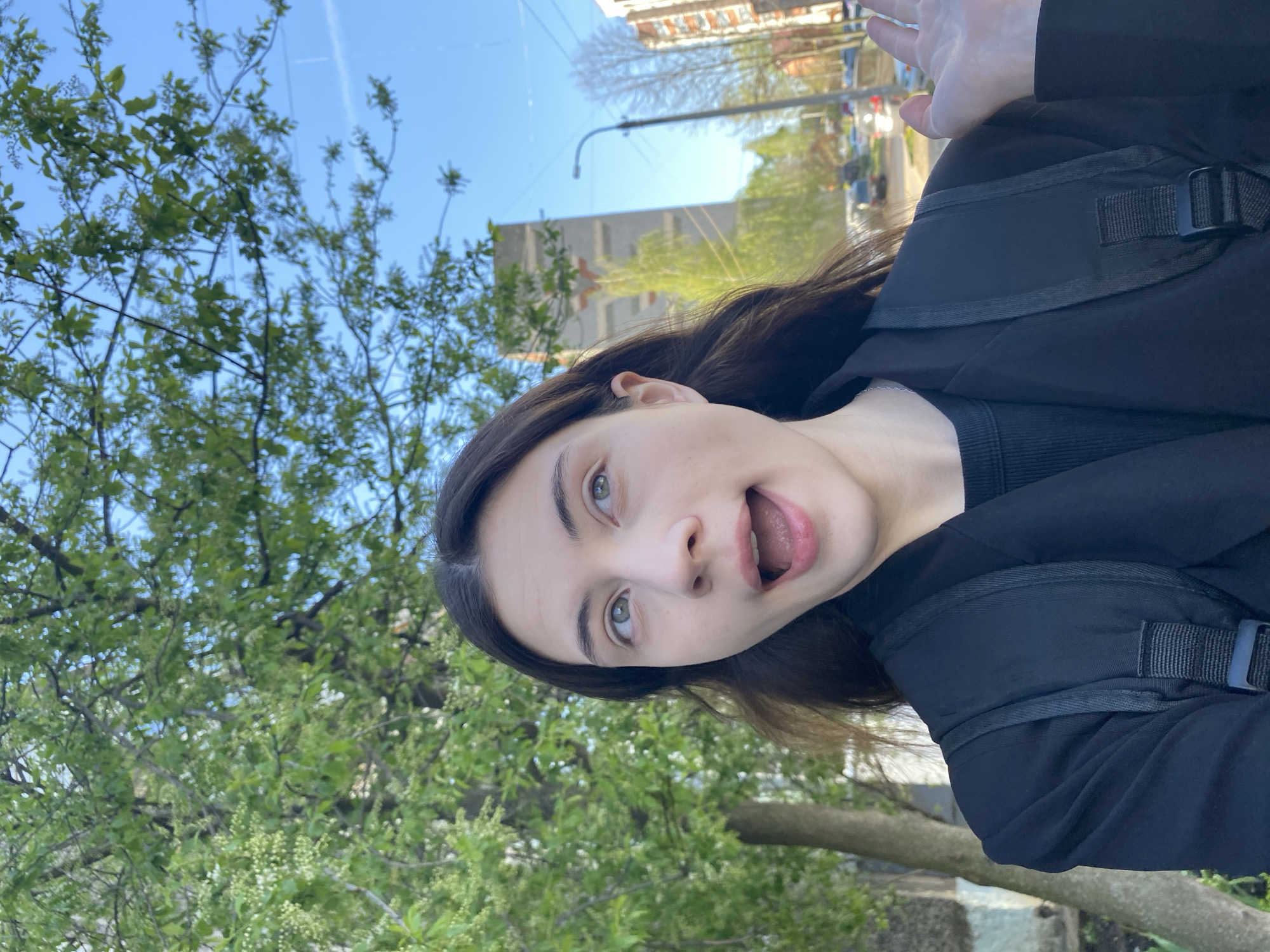 A woman is taking a selfie outdoors, likely on a street in Votkinsk, Russia. She is wearing a black jacket over a dark top and has a black backpack on. She has dark hair and fair skin. Her mouth is open, and she is looking at the camera. Her right hand is raised in a wave. The background includes greenery (trees and foliage), a building, and a street with cars. The sky is blue.