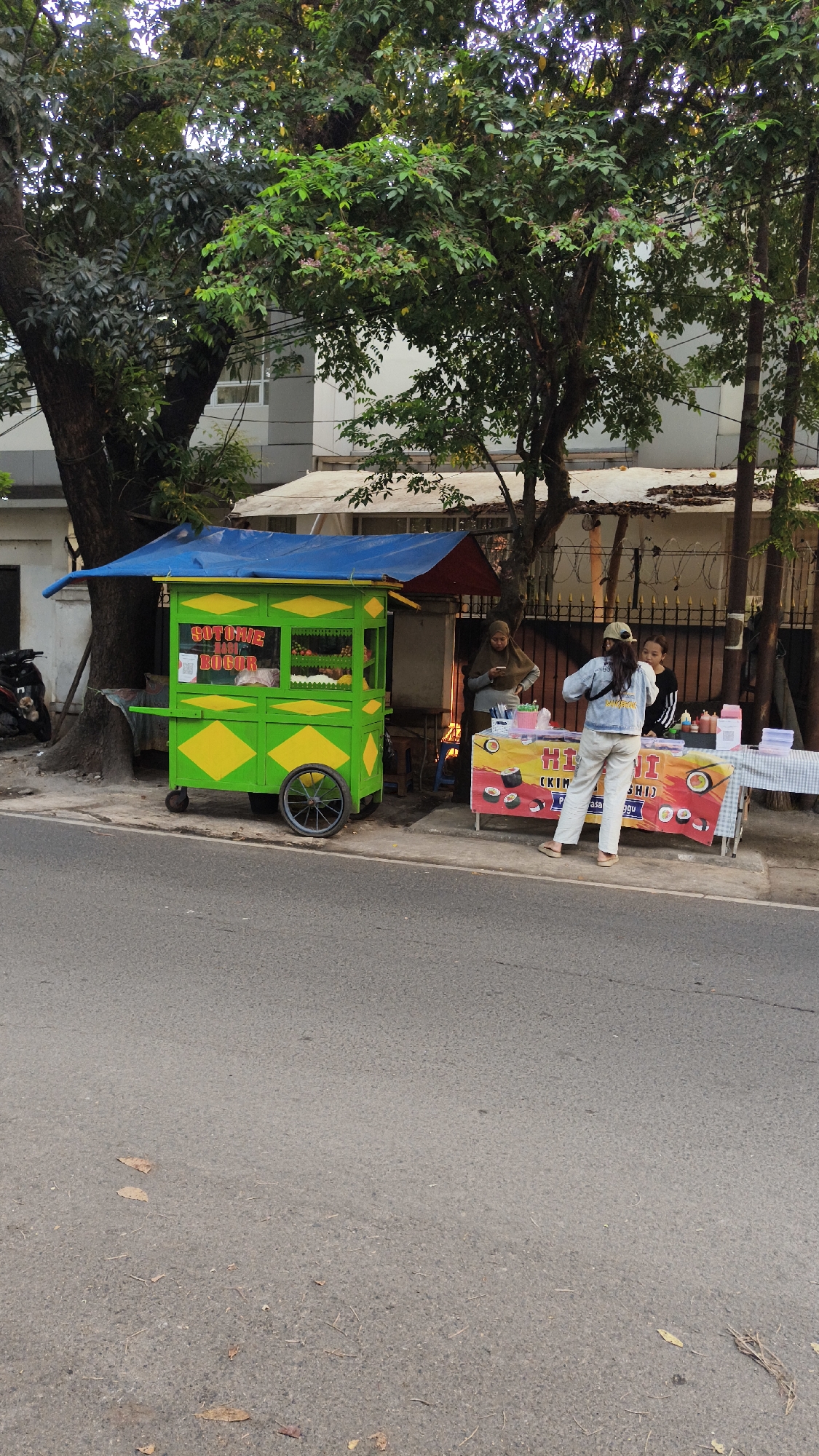 In South Jakarta, Indonesia, on a street, two food carts are set up under trees. The cart on the left is green and yellow and appears to be selling "Soto Mie Bogor." The cart on the right is covered by a banner advertising "HI NI (KIM CHI) SUSHI" and has sushi rolls displayed. A person wearing a hijab is standing behind the sushi cart, looking at their phone. Another person in a denim jacket and light-colored pants is facing the sushi cart, and a third person, possibly a vendor, is behind the sushi cart. The street is paved, and there are fallen leaves on the ground. A motorcycle is parked on the left side of the image. A building is visible in the background.