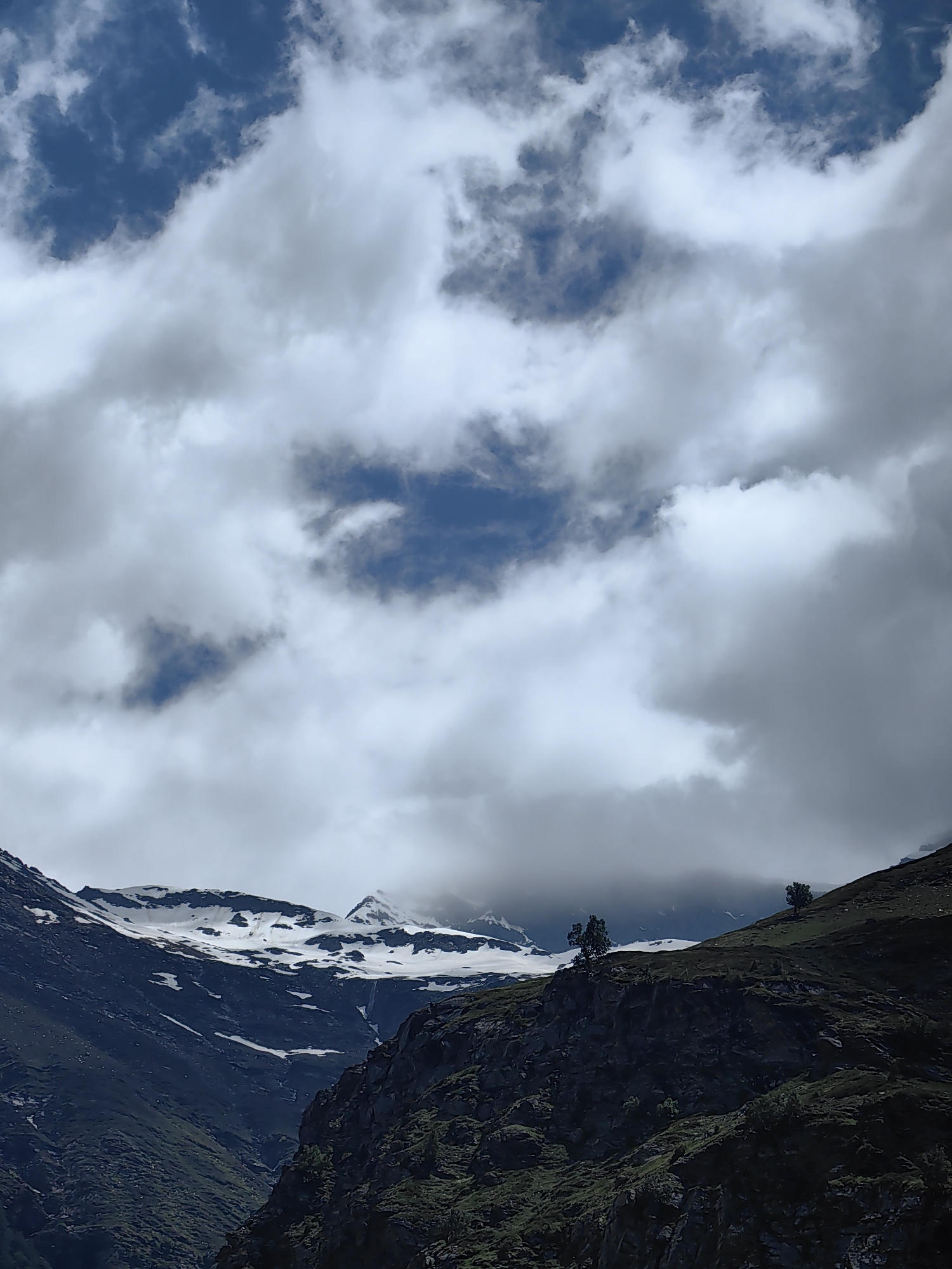 The image depicts a dramatic mountain landscape with a dynamic sky. In the foreground, dark, rocky slopes covered in green vegetation rise steeply. Two solitary trees stand on the crest of one of these hills. Beyond these slopes, towering mountains are visible, their peaks capped with snow, suggesting a high-altitude environment. The sky above is filled with a mix of dramatic, heavy clouds and patches of clear blue sky, creating a sense of depth and movement. The overall scene is one of rugged natural beauty and a vast, open environment. The location is identified as Burwa, India.
