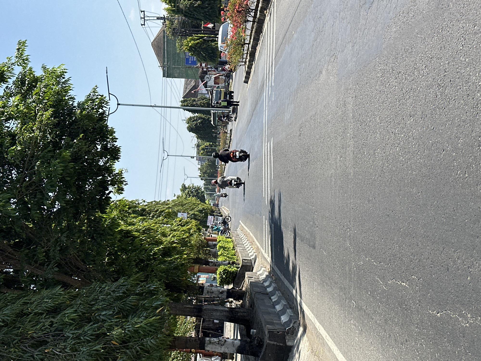 The image captures a street scene in Bulakrejo, Indonesia, on a clear, sunny day. The perspective is from a moving vehicle, looking down an asphalt road lined with trees and buildings. In the foreground, the road shows signs of wear with some cracking and patching. Several motorcycles are visible on the road, with riders wearing helmets and casual clothing. One motorcycle in the middle ground has a rider wearing a black helmet and jacket, while another rider in front is wearing a red helmet. Further ahead, a person in a yellow vest, possibly a traffic controller or worker, stands next to a truck. On the right side of the road, there are decorative shrubs and trees, and beyond them, buildings and utility poles. The left side of the road features a concrete barrier with black and white markings, lined with trees and some low-lying vegetation. A few stalls or small shops are visible along the roadside in the distance. The overall impression is of an active street in a tropical urban or semi-urban environment.