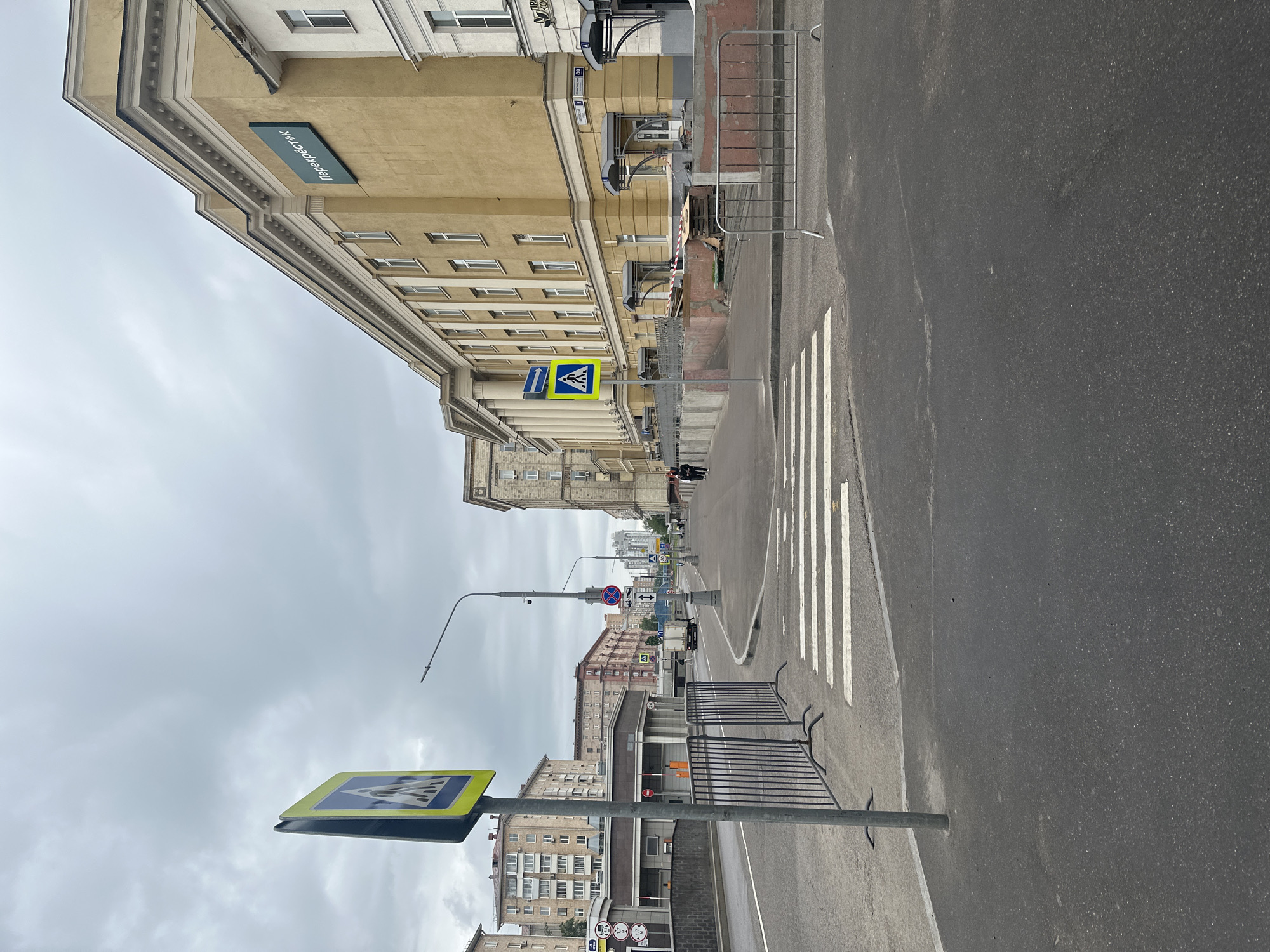 This image captures a street scene in an urban environment under an overcast sky. The primary perspective is along a multi-lane asphalt road, with a prominent pedestrian crossing marked by white zebra stripes in the foreground and mid-ground.
To the left, a large, multi-story building with a light tan or beige facade dominates the view. It features numerous rectangular windows, classic architectural details like decorative cornices and moldings, and a sign displaying the Cyrillic word "Перекресток" (Perekrestok), indicating a supermarket. Further down the street, other residential and commercial buildings are visible, some appearing older with balconies.
Along the right side of the road, metal crowd barriers or fences are set up, separating the sidewalk area from the street. A tall street lamp post with a curved arm is positioned in the middle distance. Several road signs are present: a large yellow and blue diamond-shaped sign indicating a pedestrian crossing is in the lower left foreground, and another similar sign is visible further down. A blue rectangular sign with a white arrow pointing straight ahead can also be seen. In the distance, other signs include a red circle with a diagonal line (often indicating 'No Entry' or 'No Stopping') and directional arrows.
A few small figures of people can be vaguely discerned walking on the sidewalks in the distance, but no specific activity or event is clearly depicted. The time of day appears to be daytime, given the general illumination, and the cloudy sky suggests a mild, possibly dull weather condition. The architecture and Cyrillic text on the signage are consistent with the provided location context of Khimki, Russia.