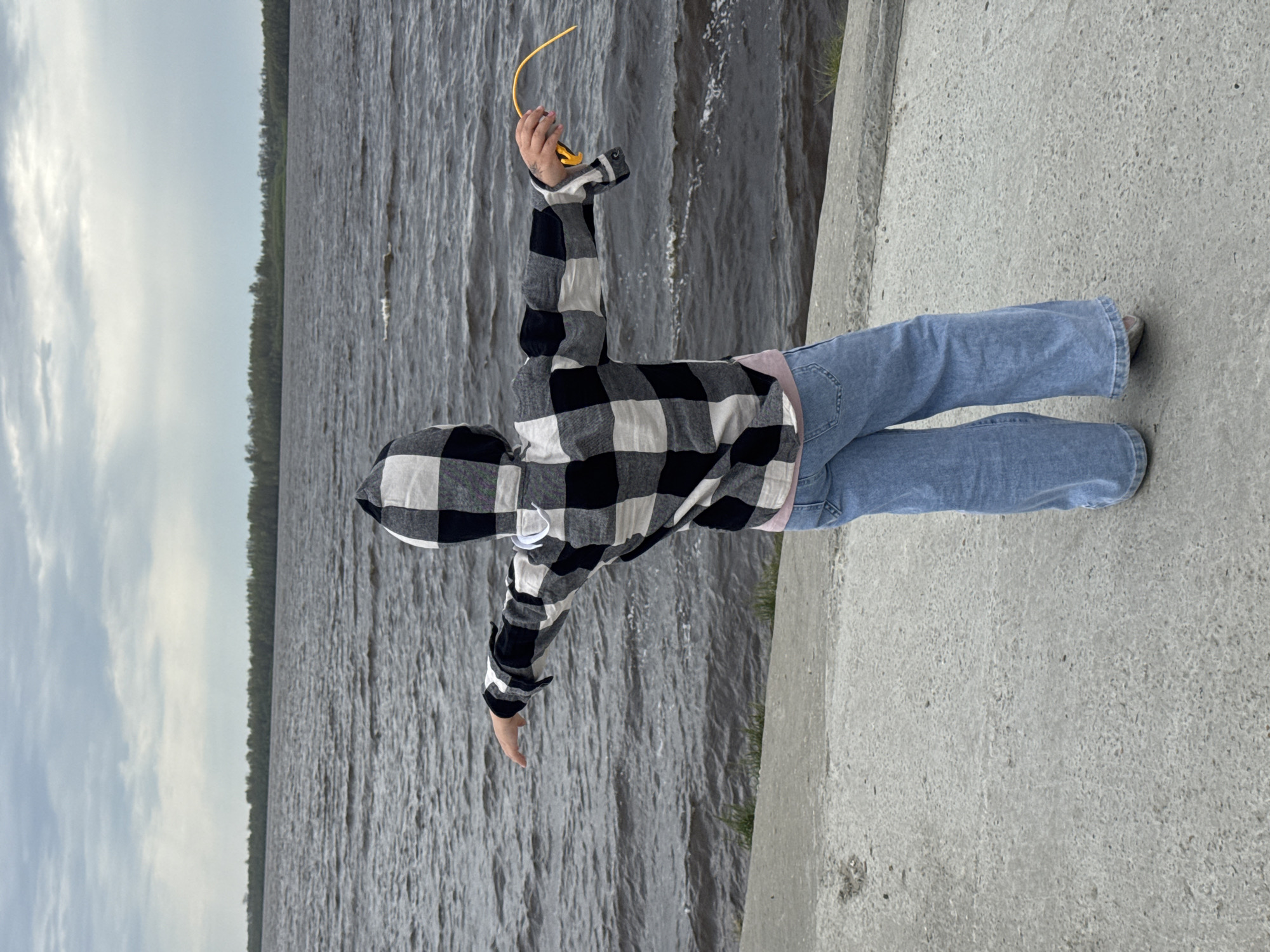 A person wearing a black and white checkered hooded jacket and blue jeans is standing on a concrete surface next to a body of water. The person is holding an object with a yellow curved end in their right hand. Trees are visible in the background across the water.
I lack the ability to determine the specific location.