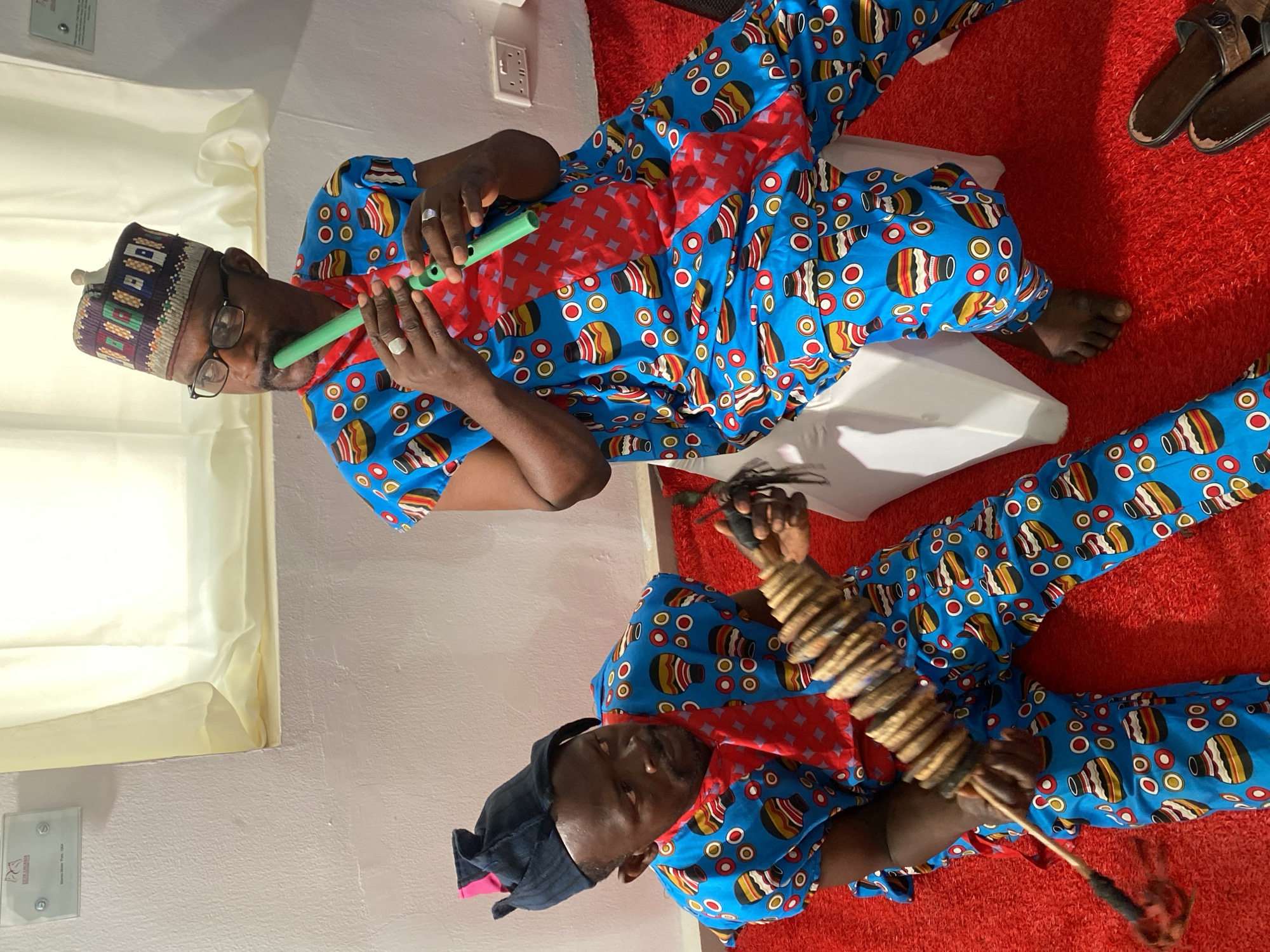 The image shows two men on a red carpet, possibly indoors. Both are wearing matching blue outfits with a pattern of colorful pots or jars and a red diamond-patterned scarf. One man is playing a green flute, while the other is holding and playing a percussion instrument. A pair of sandals is visible in the upper right corner. The location is not possible to determine from the information provided in the image, and the city of Kwarau is not mentioned.