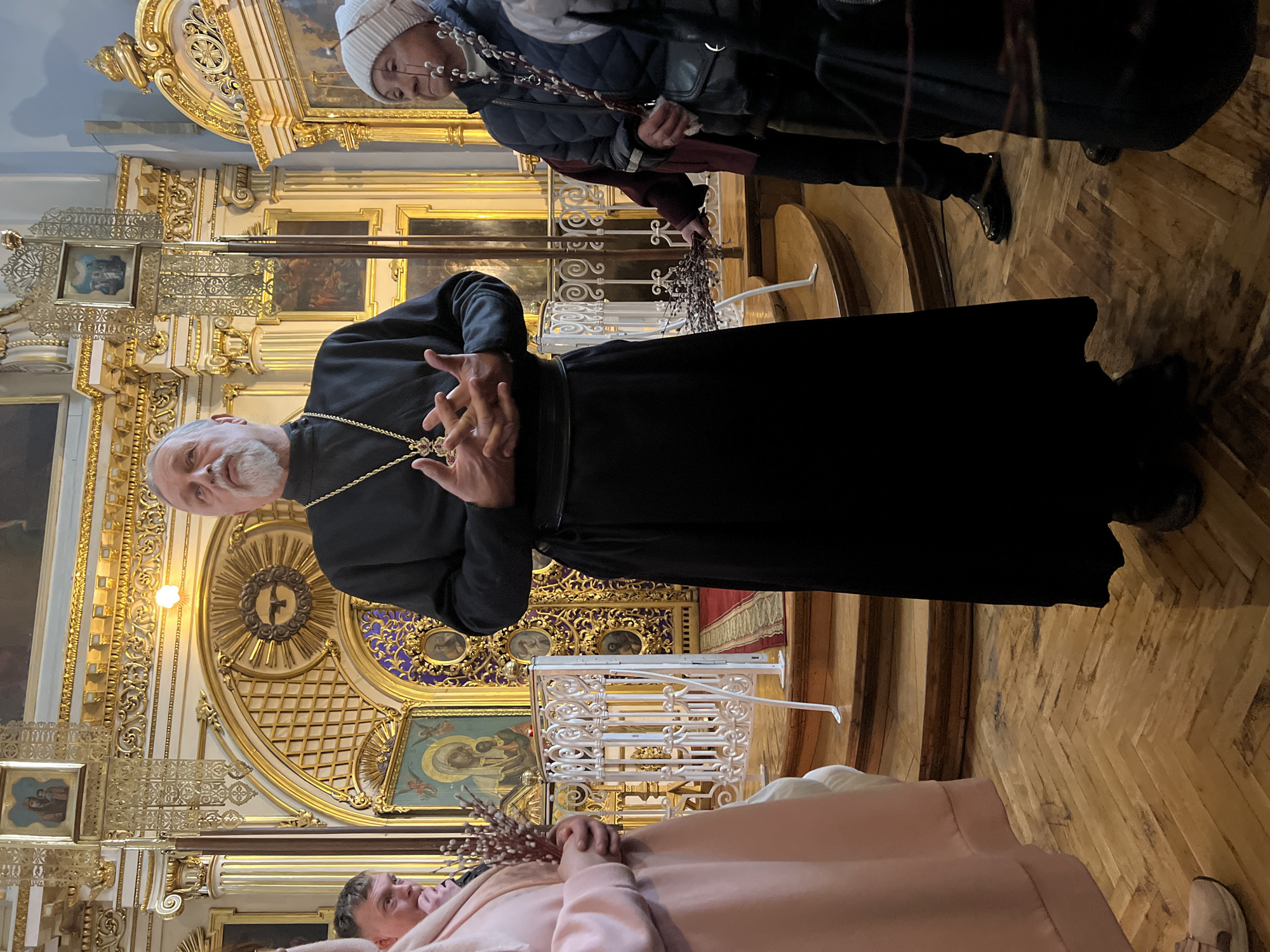 Here is a detailed description of the image:
The image shows a scene inside what appears to be an Eastern Orthodox church, likely located in Saint Petersburg, Russia, based on the architectural style and religious symbolism.
A bearded priest in a black robe and gold chain stands prominently in the frame. He has his hands together in a gesture of blessing or teaching. Behind him is the ornate gilded iconostasis, a wall of icons and religious paintings separating the nave from the sanctuary. The iconostasis is adorned with golden details, including icons of religious figures.
In the foreground, a person in a pink robe lies prone, holding a bouquet of pussy willows. This may indicate a ritual or blessing involving the priest and the person. Above the priest, other people are observing the scene, wearing regular clothes.
The interior of the church features wooden flooring, possibly parquet, and is illuminated with warm light, highlighting the golden details of the iconostasis. A white, metal railing can be seen, presumably to keep people separate from the altar.
