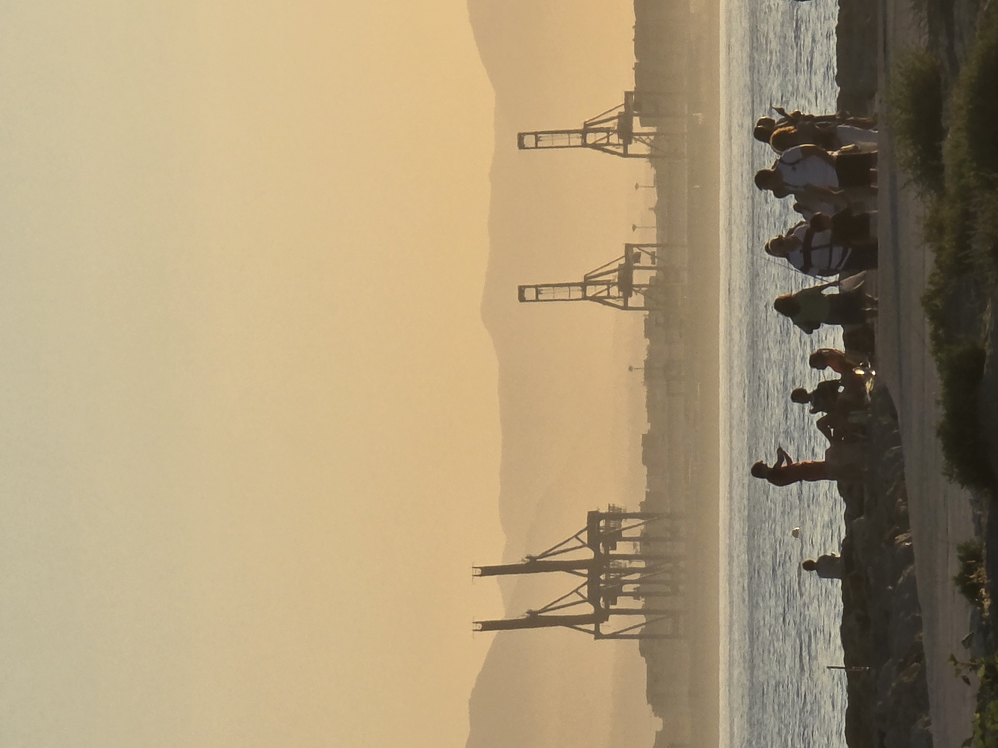 The image captures a sunset scene at the port of Malaga, Spain. In the foreground, a group of people are gathered on a rocky shoreline, silhouetted against the golden light. Some are standing, while others are seated. The sea is calm, with gentle waves lapping at the shore.
In the middle ground, the port of Malaga is visible, with several large cranes standing tall against the hazy horizon. These cranes are industrial in nature and suggest activity within the port. The background is dominated by a series of mountains, softened by the atmospheric haze and the warm hues of the sunset.
The overall mood of the image is tranquil and serene, with the fading light casting a soft glow over the landscape. The presence of people suggests a leisurely outing, perhaps enjoying the end of the day by the sea. The juxtaposition of the natural landscape with the industrial port structures creates a unique visual dynamic.