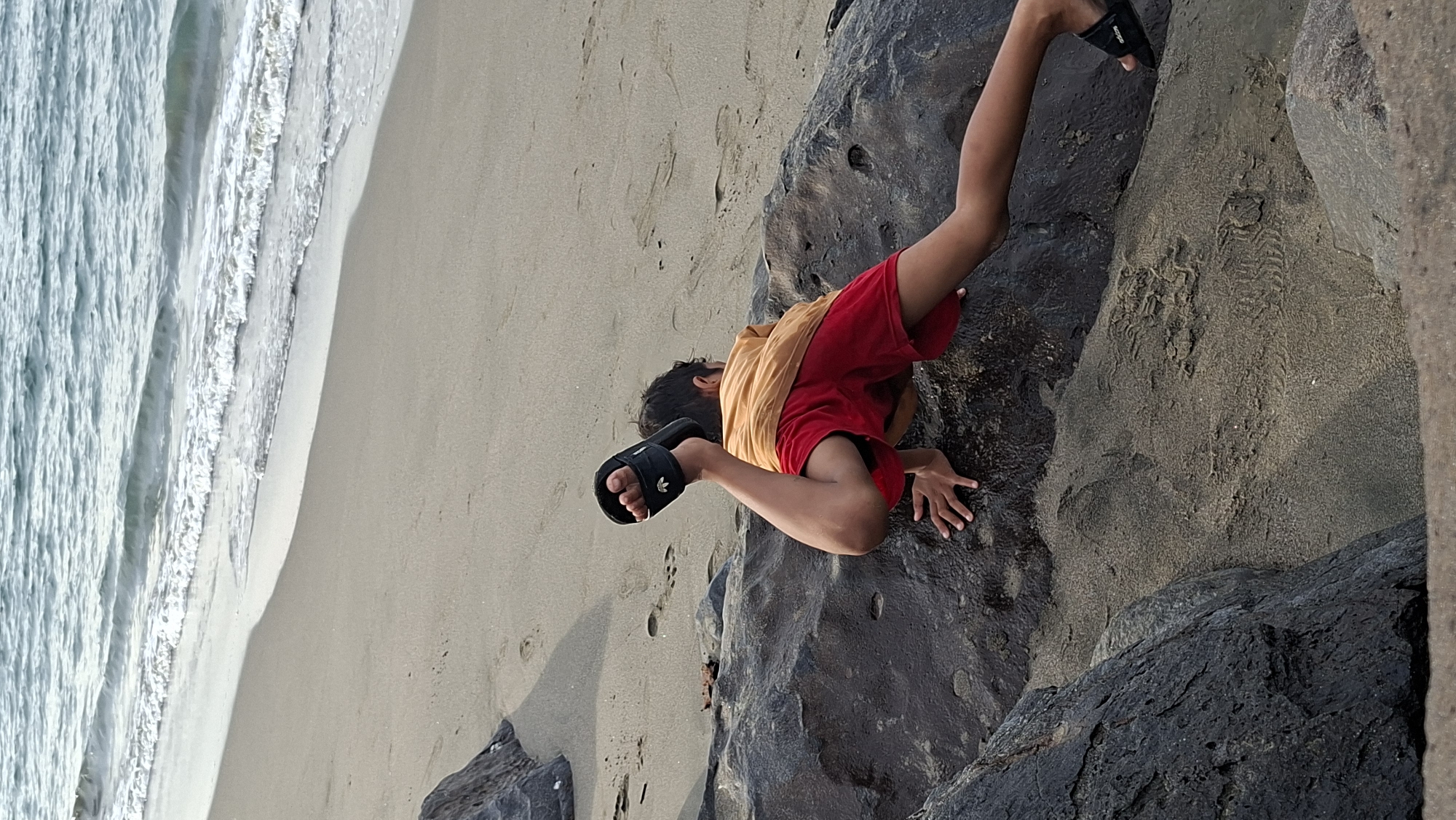 A young boy is seen on a sandy beach near Lhokseumawe, Indonesia, in the process of climbing over a dark-colored rock formation. He is wearing a faded orange t-shirt and red shorts, along with black slide sandals. His body is contorted as he maneuvers over the rock, with one leg extended and the other bent. The beach appears to be a mix of sand and scattered rocks, with gentle waves lapping at the shore in the background. The lighting suggests it may be late afternoon or early morning.