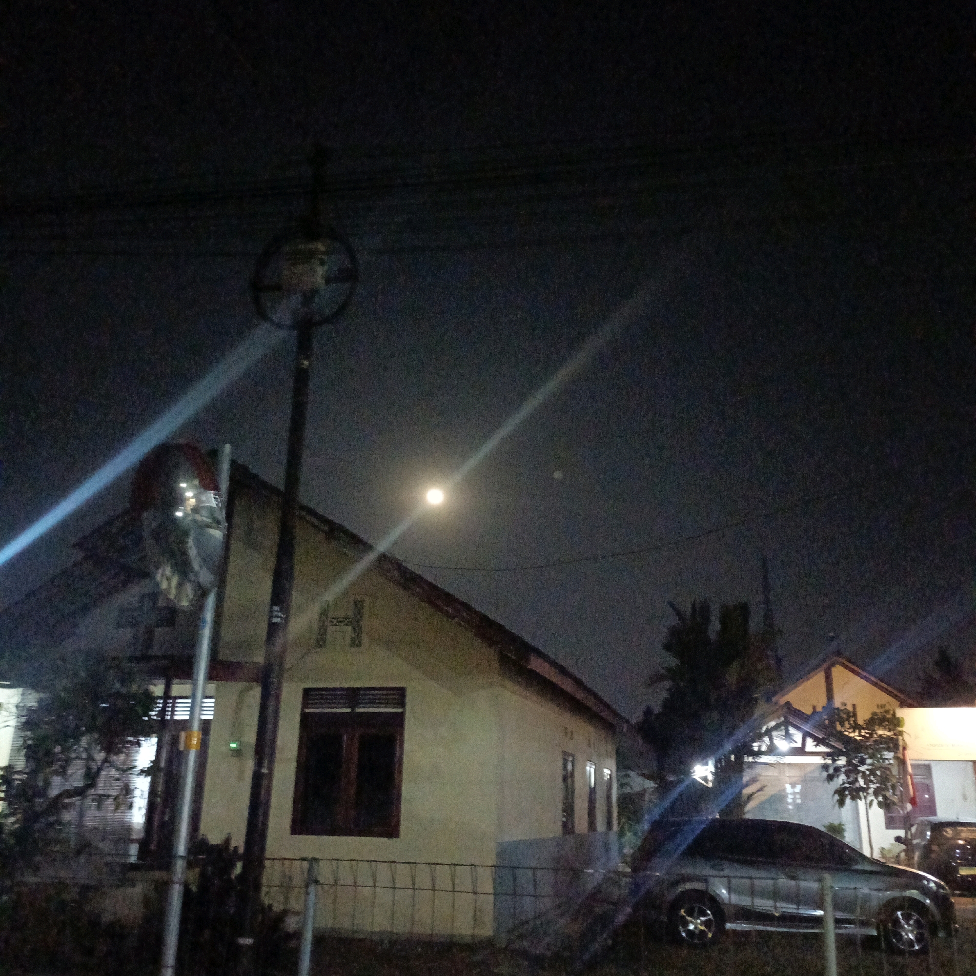 The image depicts a nighttime scene in Salatiga, Indonesia, characterized by a dimly lit sky with a visible full moon. In the foreground, a silver sedan is parked on the right side of the image, its headlights off. Behind the car, a light-colored house with a brown-tiled roof is partially visible. A street light with a circular reflector is located to the left of the house, casting a bright beam across the scene. The house has dark-framed windows and a large letter "H" appears on its facade. A metal fence runs along the front of the property. Trees and foliage are present, some appearing silhouetted against the night sky. Another building, with a white facade and a light-colored roof, is situated to the right of the primary house, with its windows illuminated. The overall atmosphere is one of quiet domesticity under the nocturnal sky.